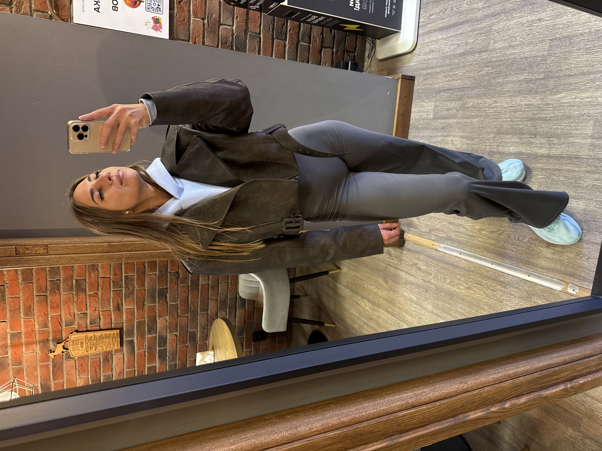 A woman is reflected in a mirror. She holds a cell phone up with her left hand, taking the photograph. She wears a light blue shirt under a brown jacket, gray pants and light blue shoes. The environment includes a brick wall, a gray wall and a gray wooden floor. A chair sits behind her. A black poster is visible in the upper part of the photograph. The photograph appears to be taken in Saint Petersburg, Russia.