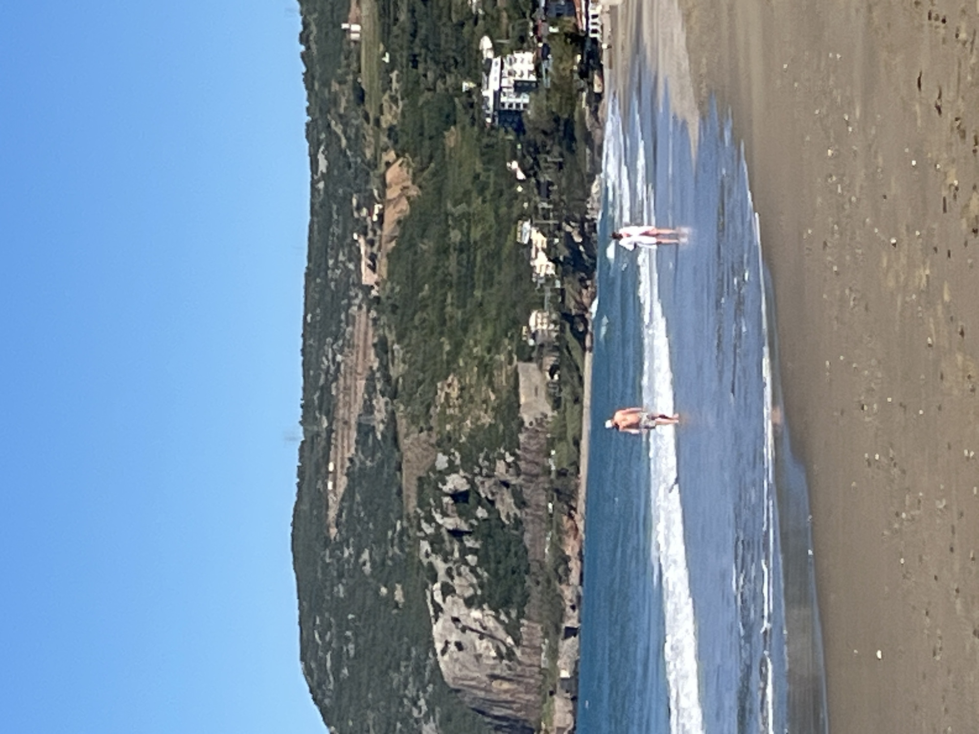 The image shows a beach scene in Alanya, Turkey. The view is a slightly rotated, ground-level shot. In the foreground, a sandy beach transitions into the shoreline. The water is blue with white foamy waves. Two people are visible in the water, one further away and one closer to the camera. A large, green, rocky hill with buildings dotted on it rises up in the background. The sky above is clear and blue.