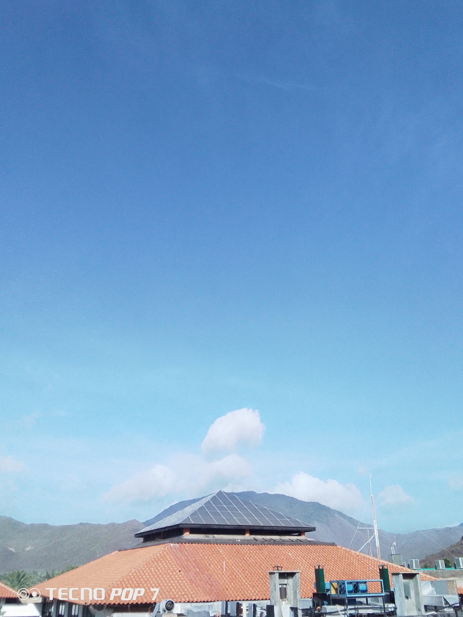 The image shows a cityscape in Pedro González, Venezuela, under a clear blue sky. The foreground is dominated by a terracotta-tiled roof with a modern addition: a pyramid-shaped structure covered in solar panels. Behind this structure are rolling, green hills. A radio antenna is visible in the distance. Various mechanical equipment, including what appear to be air conditioning units, are situated on top of the roof in the middle ground. The sky is a gradient of blue. The image was taken with a Tecno Pop 7 phone, as indicated by the watermark in the lower left corner.