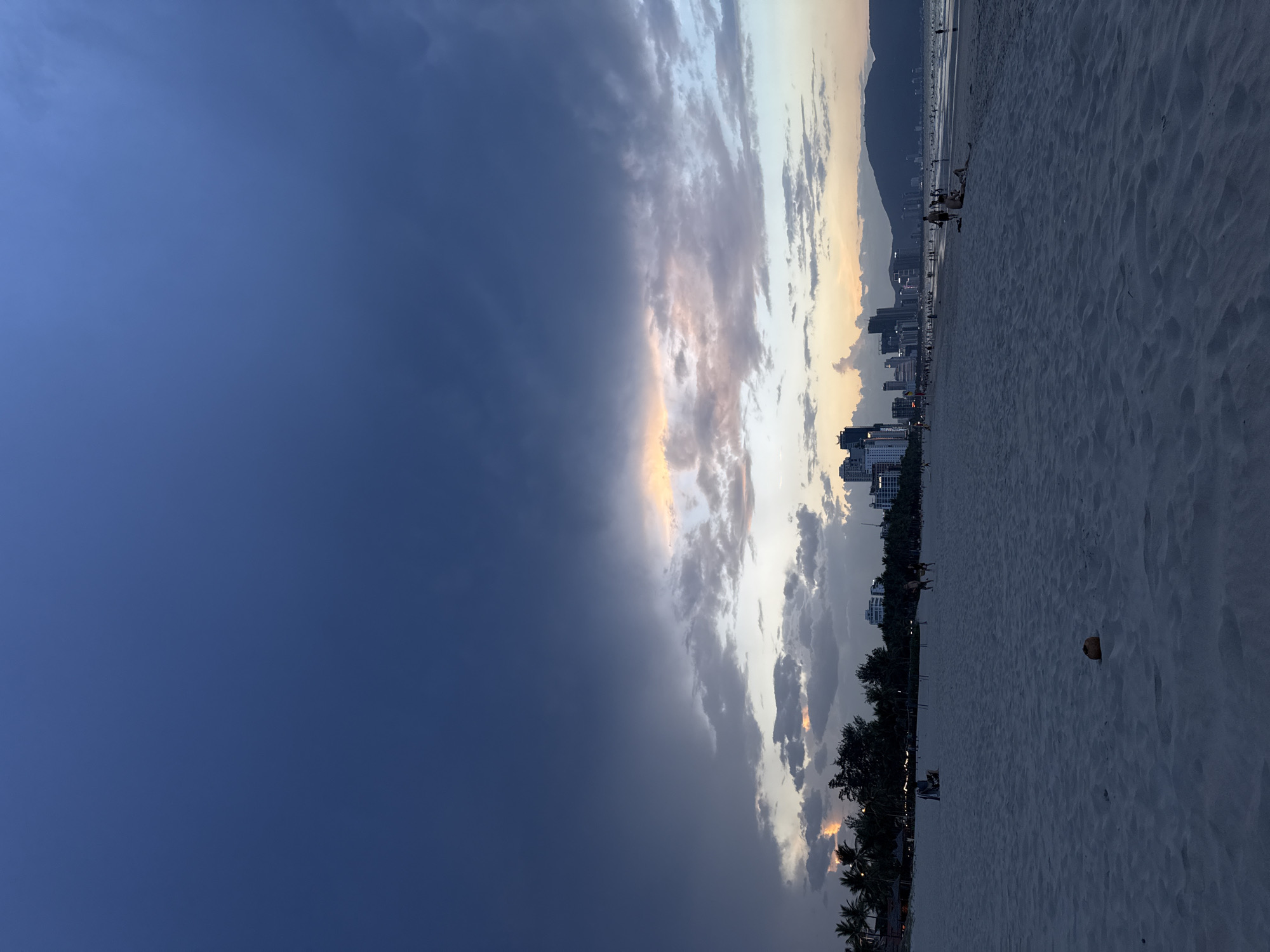The image captures a panoramic view of a beach in Đà Nẵng, Vietnam, at what appears to be dusk or dawn. The sky is a prominent feature, dominated by a large expanse of dark blue-grey clouds on the left, transitioning to lighter, scattered clouds with visible hues of orange and yellow on the right, suggesting a sunset.
Along the horizon, a city skyline with various buildings is discernible, backed by distant mountains silhouetted against the lighter sky. The foreground is a wide, light-colored sandy beach exhibiting ripples and textures. Scattered individuals, appearing as small, dark figures, are present on the sand, engaged in leisurely activities such as walking or relaxing. A small, dark, round object, possibly a coconut, lies on the sand in the mid-ground. A line of trees, including palm trees, borders the beach, enhancing the tropical setting.
The scene conveys a peaceful yet dramatic atmosphere due to the contrasting cloud formations and the interplay of natural light. No visible text is present in the image.