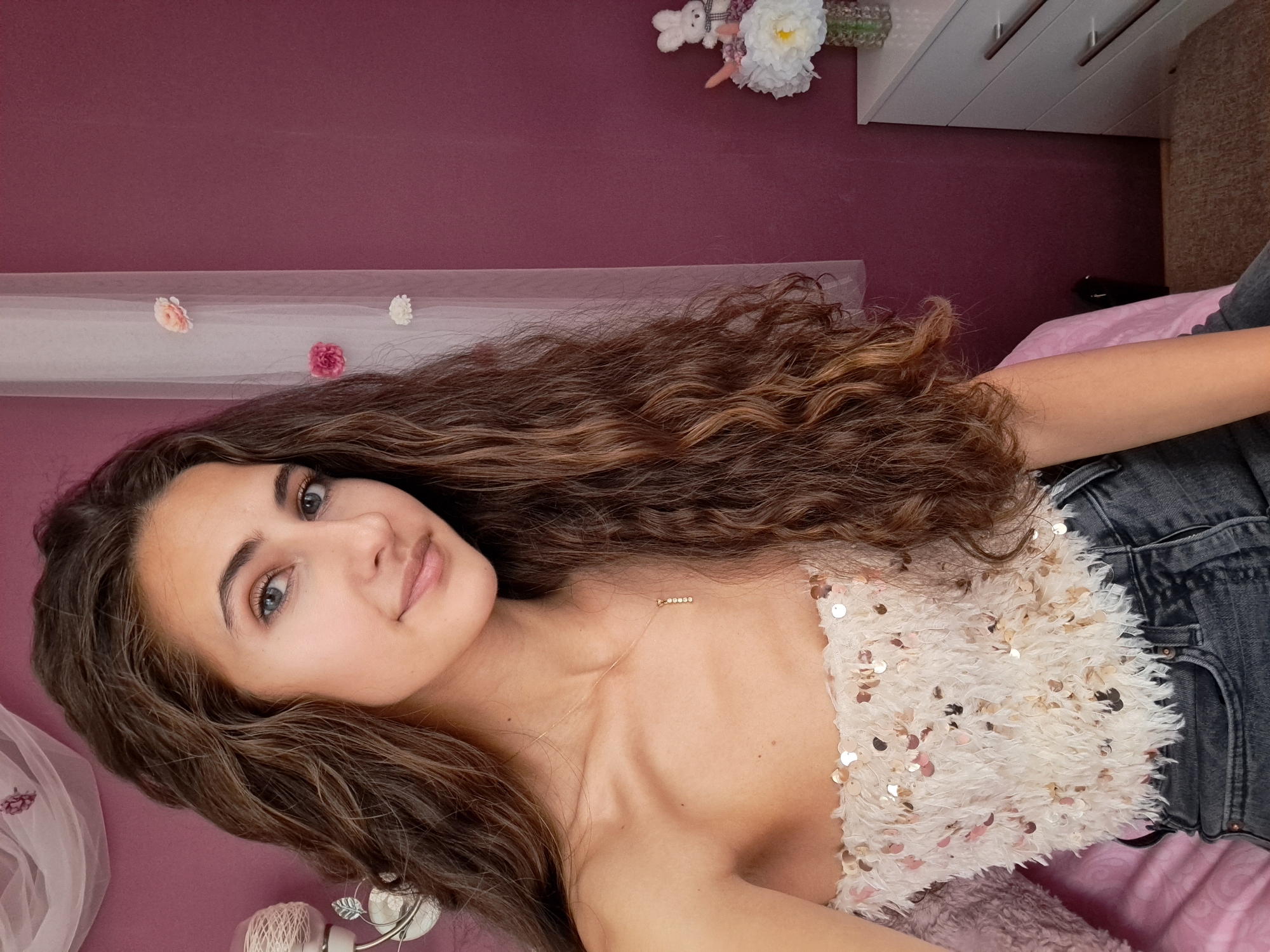 A young adult female is depicted seated indoors in what appears to be a bedroom, looking directly at the viewer with a slight, closed-mouth smile. Her long, wavy to curly hair, dark brown with lighter auburn or caramel highlights, cascades over her shoulders. She has blue-grey irises and appears to be wearing light makeup. She is dressed in a strapless, off-the-shoulder top composed of a shaggy, light-colored fabric interwoven with numerous small, iridescent sequins, paired with dark grey high-waisted denim jeans. A thin gold-colored chain necklace with a small pendant featuring a vertical line of five clear, possibly diamond or crystal, stones rests on her décolletage.
The background features a deep plum, magenta, or berry-colored wall. To the left, an ornate light-colored wall sconce with a decorative leaf motif holds a spherical light fixture wrapped in a textile material. Above it, sheer white fabric, possibly part of a bed canopy or curtain, is draped. To the right, a sheer white curtain hangs vertically, adorned with three vertically spaced fabric flowers: one pale pink, one deep pink, and one white. Further right, a white three-drawer dresser with horizontal silver-colored pull handles is visible. On top of the dresser sits a small white plush bunny toy with a black and white checkered bow, alongside a small clear glass vase containing artificial white and light pink floral arrangements. The woman is seated on a bed, partially covered by a pink sheet or blanket. A light brown carpet is visible on the floor in the bottom right corner. The location is in the city of Unknown, Unknown.