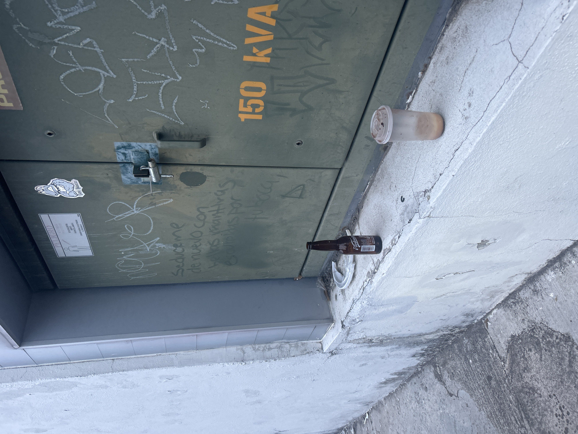 The image shows the side of a green electrical box, likely located in Puebla, Mexico, judging by the Spanish graffiti. The box is covered in graffiti, including the number "150 kVA," and is secured with a latch. A sticker is also visible on the box. Lying on the concrete surface adjacent to the box are two pieces of litter: an empty plastic cup with remnants of a brown substance inside, and an empty brown glass beer bottle, possibly Corona. A white plastic fork or spoon is also visible near the bottle. The surrounding area is a concrete surface with signs of wear and weathering.