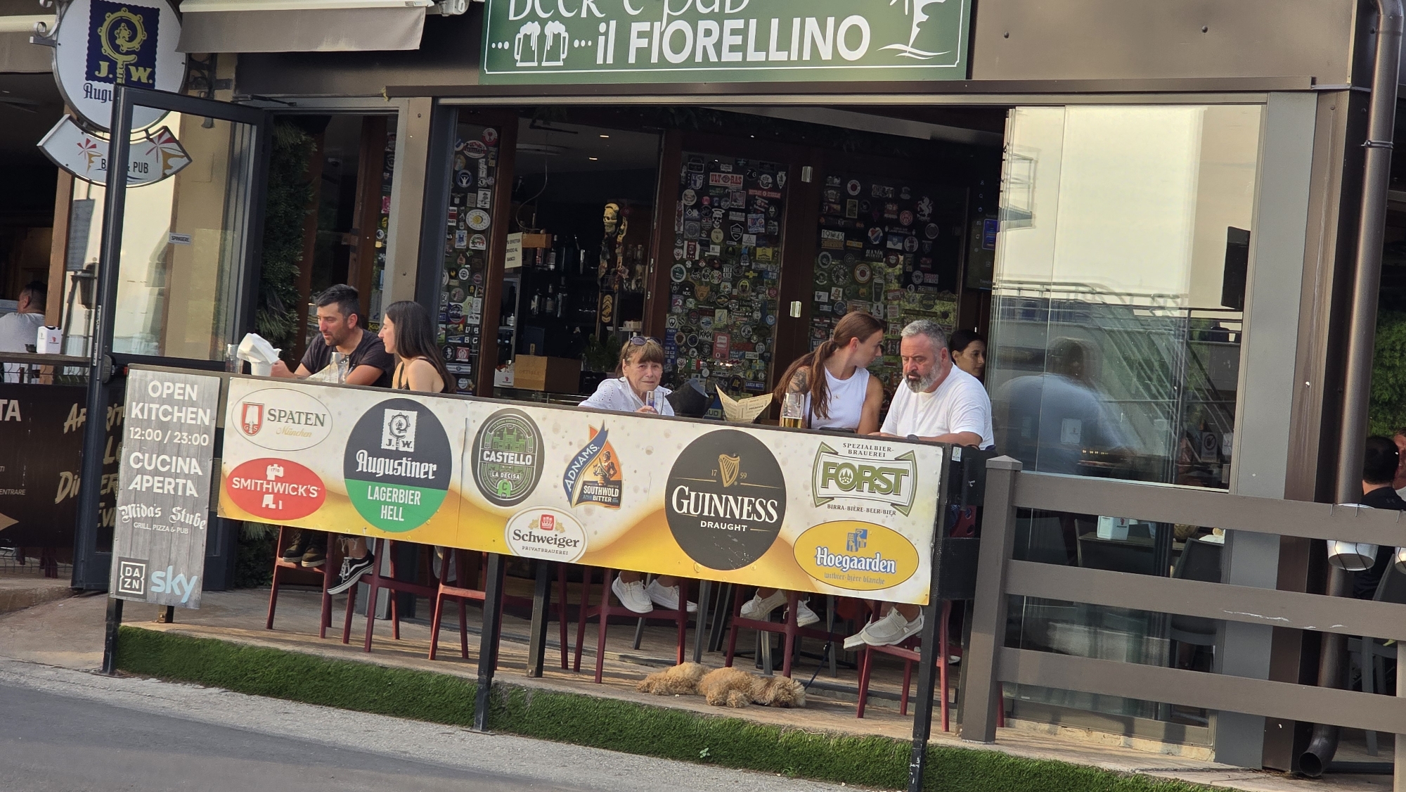 This media file captures a lively outdoor seating area of a pub called "il FIORELLINO" in Peschiera del Garda, Italy. The scene is set during daylight, likely late afternoon or early evening, indicated by the warm, golden light. Several people are seated at tables, some engaged in conversation, others appearing to dine or drink.
The pub's facade is decorated with numerous stickers, hinting at a vibrant and well-traveled clientele. Prominent signs display "OPEN KITCHEN 12:00/23:00" and "CUCINA APERTA," along with the pub's name, "il FIORELLINO." Advertising banners showcase various beer brands including Spaten, Augustiner, Smithwick's, Adnams Southwold Bitter, Guinness, Forst, and Hoegaarden. Two small dogs are resting on the grass in front of the seating area.
The overall atmosphere appears relaxed and social, with patrons enjoying the pleasant weather and the offerings of the establishment. The scene is a typical representation of a European pub or beer garden, emphasizing a casual dining and drinking experience.