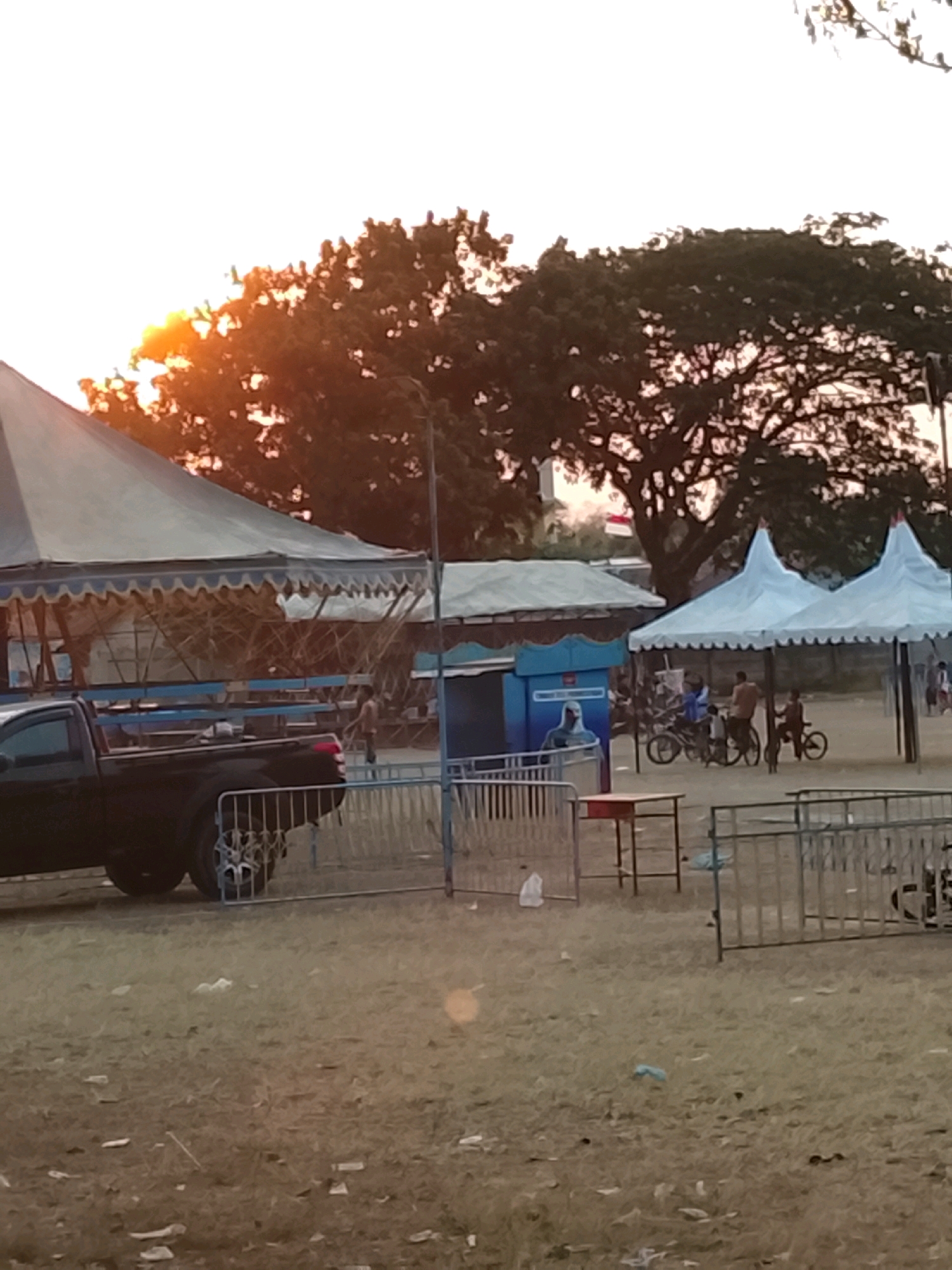The image depicts an outdoor scene at dusk, possibly in Harjosari Lor, Indonesia. A large, light-colored tent with a striped awning dominates the left side of the frame. In front of the tent, a black pickup truck is parked, facing left. A metal fence runs across the foreground.
To the right, two smaller, white peaked tents are set up. Underneath these tents, several people are visible, some with bicycles. Two individuals, one wearing a blue shirt and the other in an orange shirt, are near a bicycle. Another person, possibly a child, is seated on a bicycle to the far right of this group.
In the background, large trees with dark foliage are prominent against the bright, hazy sky, with the sun's rays filtering through the branches, casting a warm glow. A blue structure with graphics is partially visible behind the tents. The ground is covered in dry grass and scattered debris, suggesting an open field or fairground. The overall atmosphere is casual and suggests a public gathering or event in progress during the late afternoon or early evening.