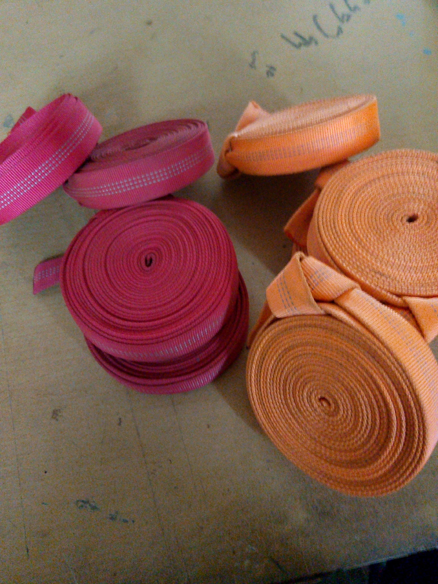 A close-up, high-angle shot shows several rolls of bright pink and orange ribbon or webbing laid out on a speckled tan surface. The pink rolls are on the left side of the frame, with one partially unrolled. The orange rolls are on the right, also partially unrolled. The ribbon appears to have a textured pattern. Faint, illegible writing in a dark color is visible on the surface in the upper right portion of the image. There are no people or animals present. The surface resembles a tabletop or a workbench. The location in Kuripan Utara, Indonesia, is not visually discernable from the provided image.
