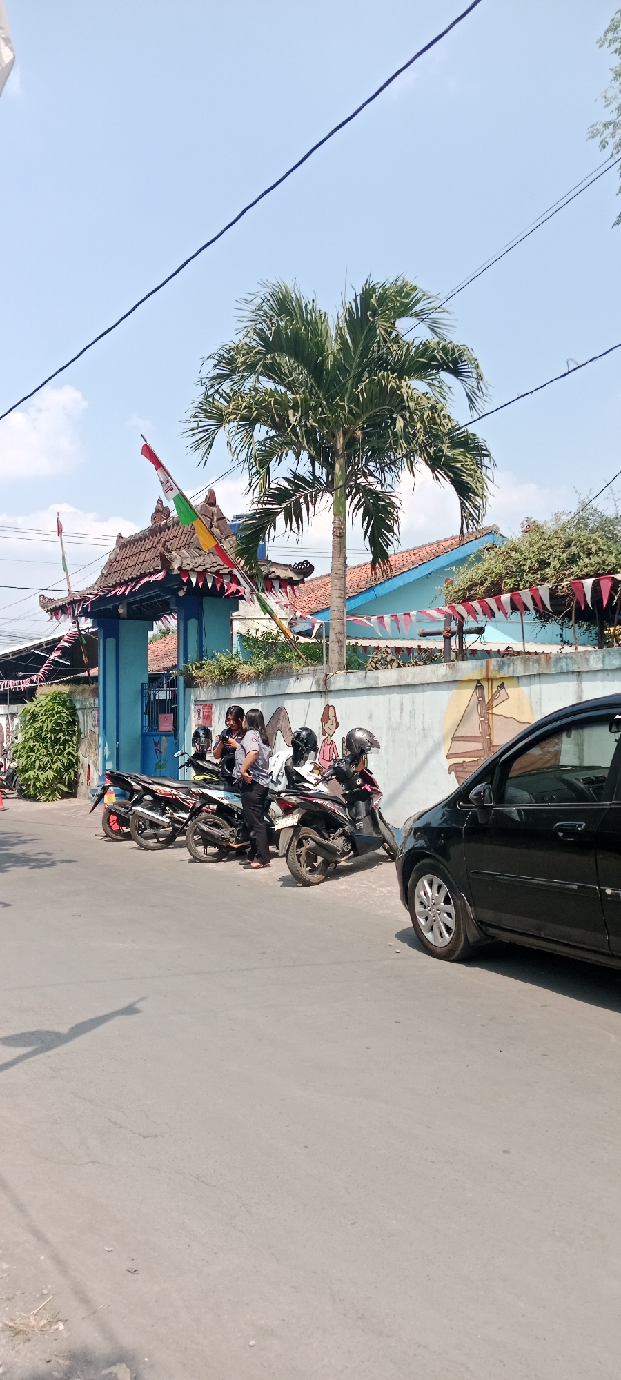 In the city of Purwomartani, Indonesia, a street scene unfolds under a clear blue sky with sparse clouds. A black car is parked on the right side of the asphalt road, its silver wheel visible. To the left, several motorcycles are parked along a light-colored concrete wall. Three individuals, two women and one person dressed in a grey uniform, are standing near the motorcycles, with one woman looking at her phone and the other two conversing. The wall itself is decorated with colorful murals depicting stylized human figures. Beyond the wall, a blue building with a red-tiled roof is visible, adorned with festive red and white pennant flags strung along its facade. A prominent palm tree stands tall in front of the building, its fronds reaching towards the sky. A traditional Indonesian gateway, also decorated with colorful flags, marks an entrance. Electrical wires are strung across the top of the frame, and the shadow of a pole or sign stretches across the road in the foreground. The environment suggests a warm, sunny day, possibly during a local celebration or holiday.