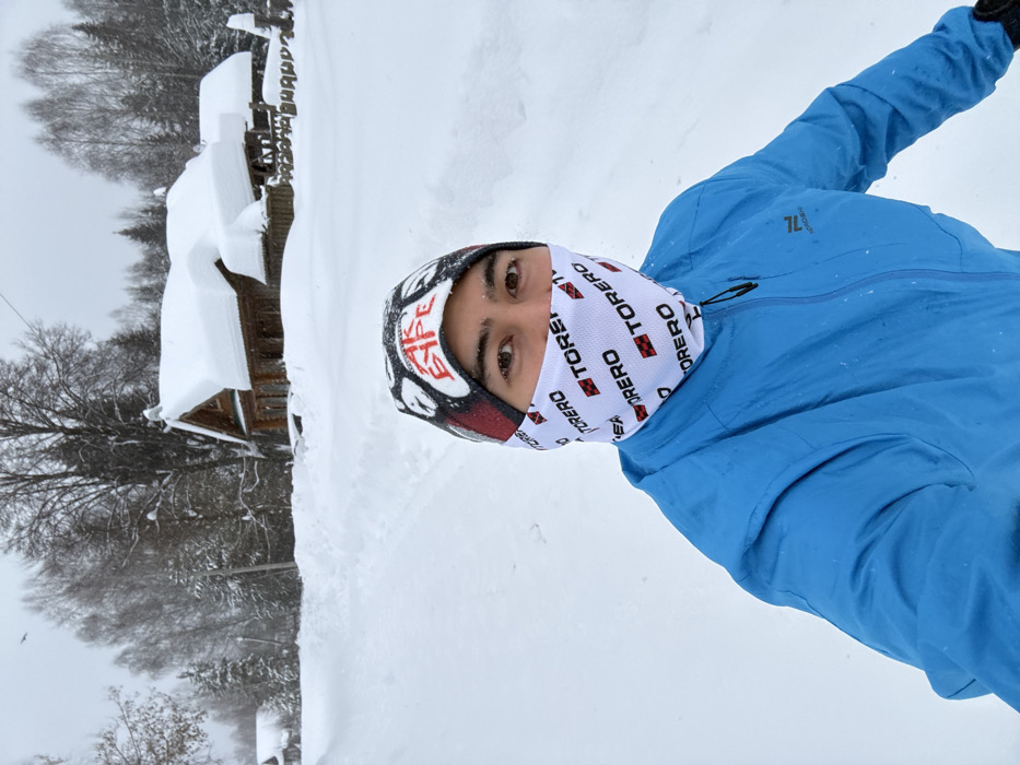 A person in a blue jacket and a balaclava stands in snowy terrain, with a building covered in snow visible in the background. The person's face is partially covered, leaving only their eyes visible. Snow covers the ground and surrounding landscape. The overall scene suggests a cold, wintery environment.