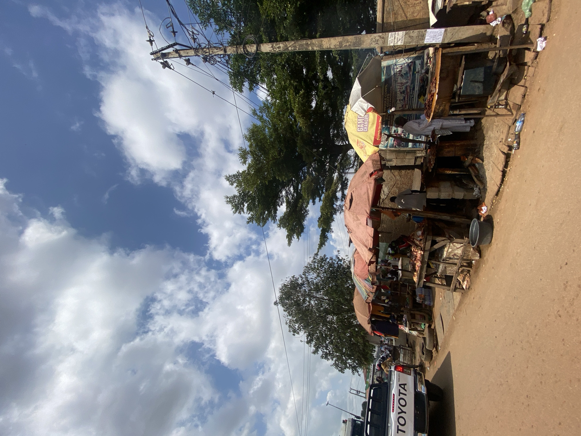 The image depicts a street scene in Ado, Nigeria, featuring an outdoor market. In the foreground, a portion of a white Toyota pickup truck is visible on the left side. The street is paved with reddish-brown dirt. A series of market stalls are set up along the side of the road, covered by various colored umbrellas. Some stalls display goods, including what appears to be meat on wooden tables, and other items hang from racks. Several people are present, some tending to the stalls and others possibly shopping or passing by. One stall has a yellow umbrella with text that reads "GAME CHANGER" and "Upgrade Your Fridge." In the background, lush green trees rise above the market stalls. A utility pole with numerous wires is prominently positioned on the right side of the image. The sky is blue with scattered white clouds. The overall impression is of a bustling local marketplace in Nigeria.
