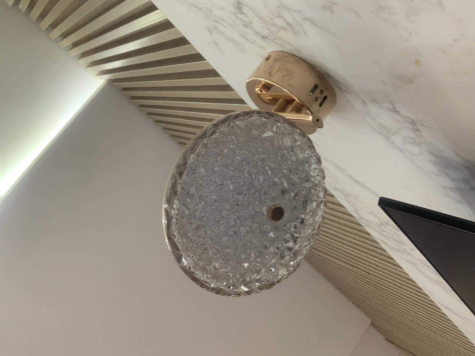 The image shows an interior shot of what appears to be a modern living space. A gold wall sconce with a decorative glass shade is mounted on a marble-patterned wall. To the left of the sconce, a portion of a flat-screen television is visible. The background features a slatted wooden wall paneling, with a linear light fixture illuminating the ceiling above. The scene is well-lit, suggesting it is daytime or the interior lighting is effective. No people or animals are present in the frame. The overall impression is one of contemporary interior design. This scene is located in Abuja, Nigeria.