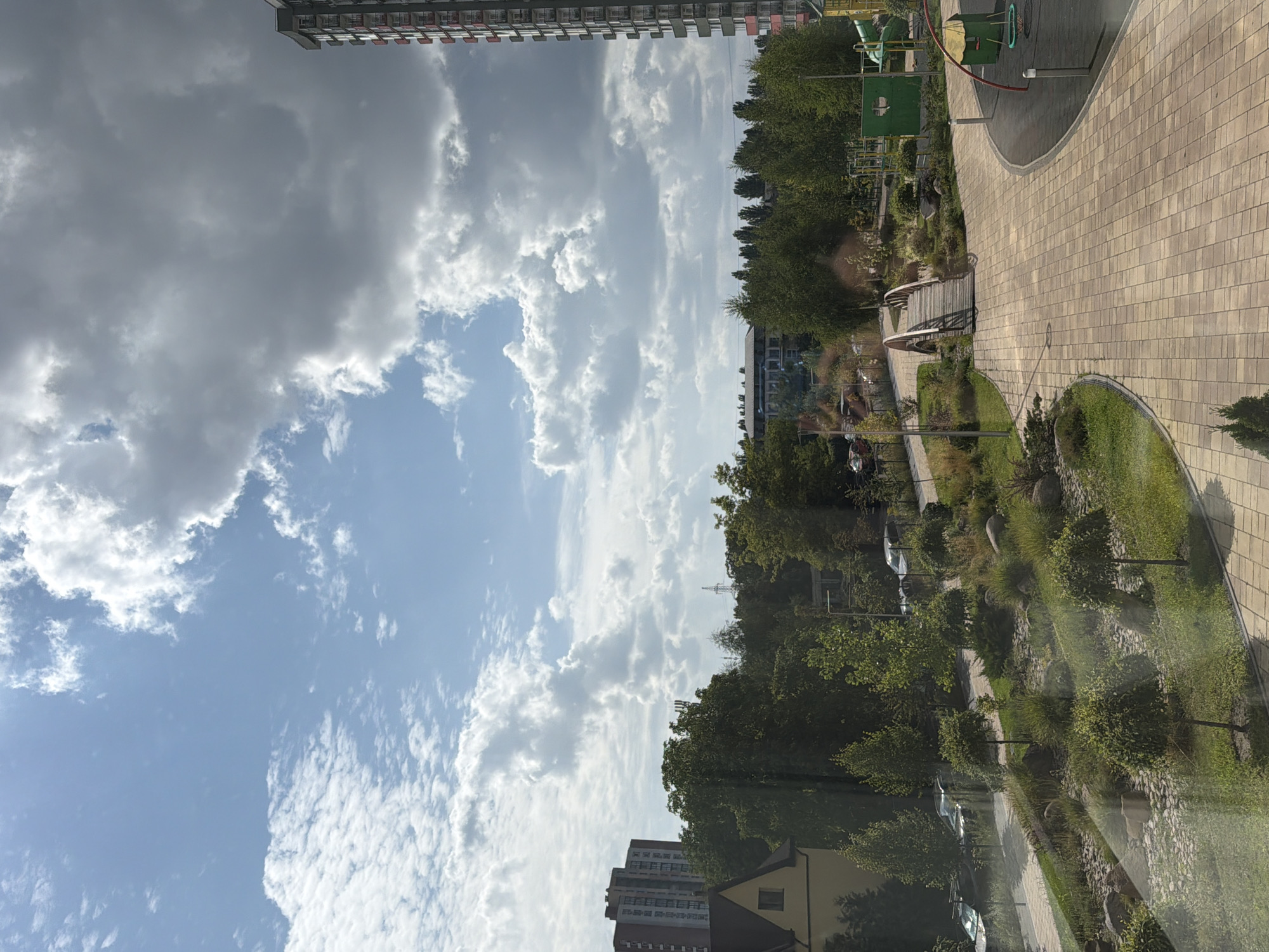 The image captures an outdoor scene in a modern residential area, likely in Kyiv, Ukraine, under a partly cloudy sky. In the foreground, a paved walkway made of light-colored rectangular tiles curves through a landscaped area with decorative grasses, small trees, and rocks. A small wooden bridge spans a dry creek bed or a decorative water feature. To the right, a playground area is visible with a green structure and red-rimmed equipment.
In the midground, a dense line of green trees separates the landscaped area from urban elements. Further back, several buildings are visible: a tall, modern apartment building with a red accent on its balconies dominates the right side of the frame. To the left, a shorter, multi-story brick building with many windows and a yellow-roofed house are present. Beyond the trees, a few cars are parked along a street. In the distance, the skyline is marked by more apartment buildings and structures, including what appears to be a cell tower and a power line pylon. The overall impression is of a well-maintained urban green space adjacent to residential development.