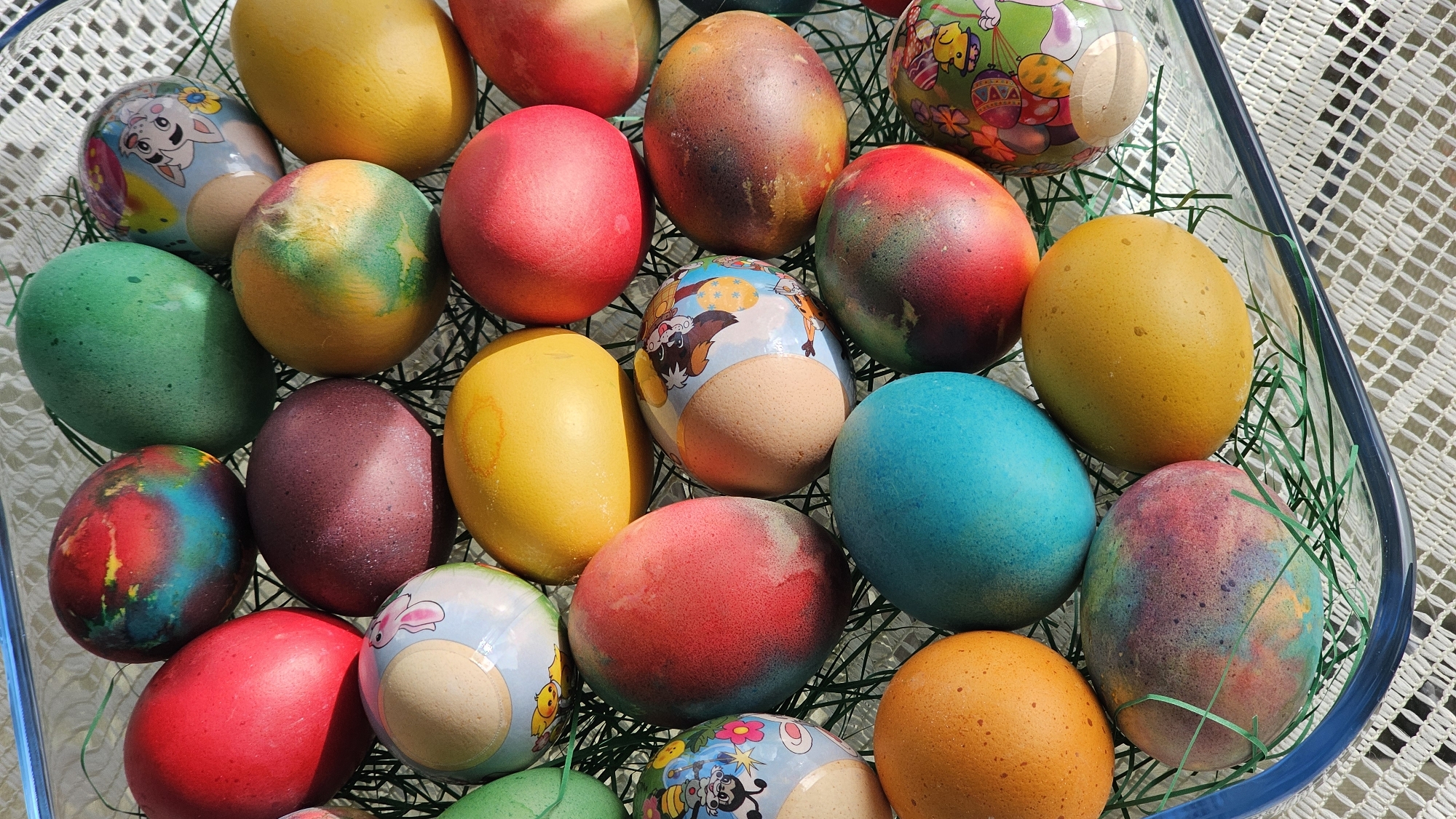 A close-up, high-angle shot depicts a glass dish filled with vibrantly colored Easter eggs nestled among artificial green grass. The eggs are various hues including red, yellow, green, blue, brown and speckled. Some are decorated with cartoon characters and patterns. The dish rests on a white, patterned tablecloth. The location is Varna, Bulgaria.
