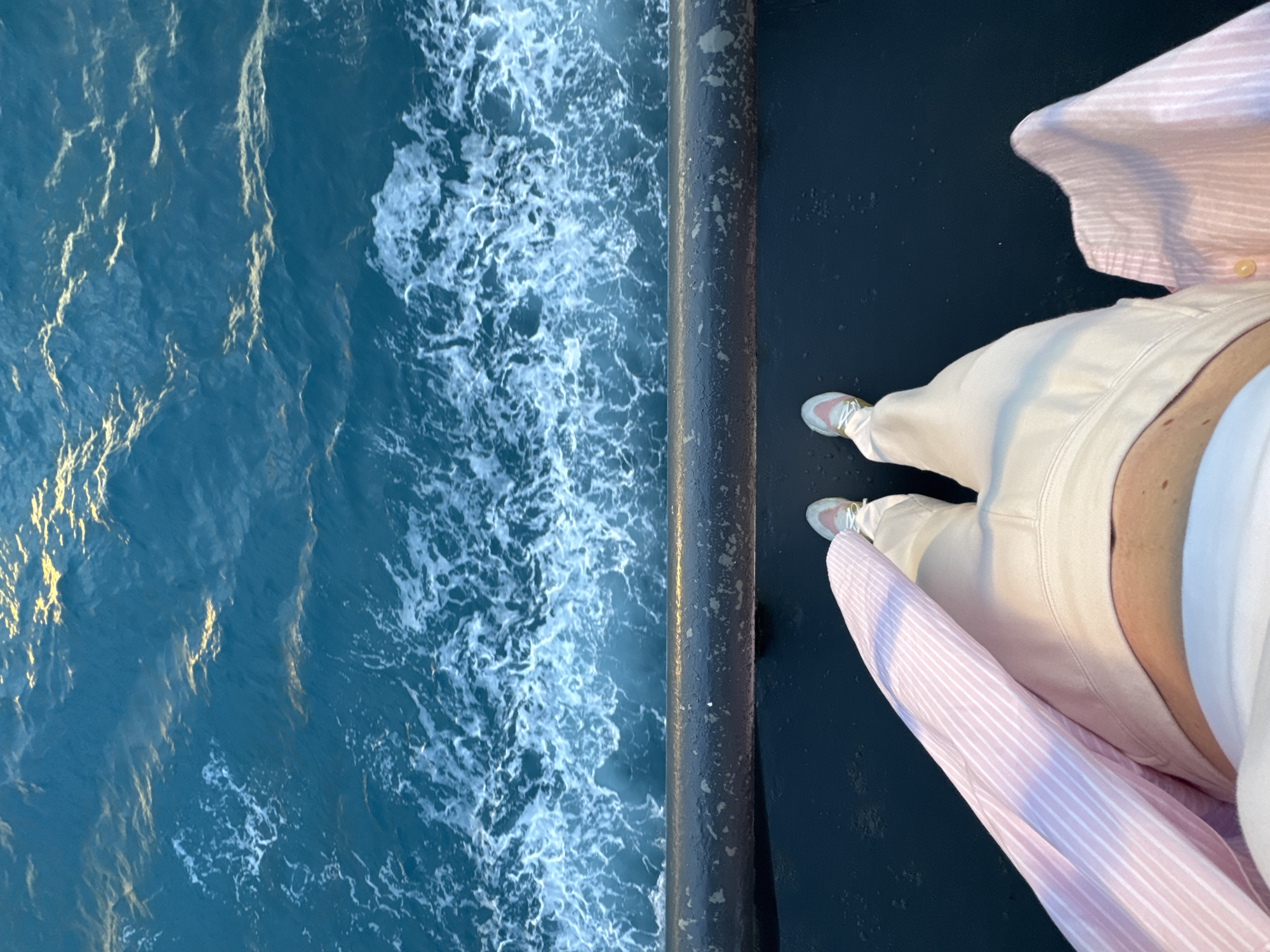 An overhead shot shows a person's legs and torso standing on a dark, flat surface, possibly a deck. The person is wearing light-colored pants and sneakers. A pink and white striped shirt is draped over their left leg. The surface they are standing on is adjacent to a dark railing, beyond which is the ocean. The water is a deep blue with white foam and waves. The sun is reflecting off the water, creating streaks of yellow light.