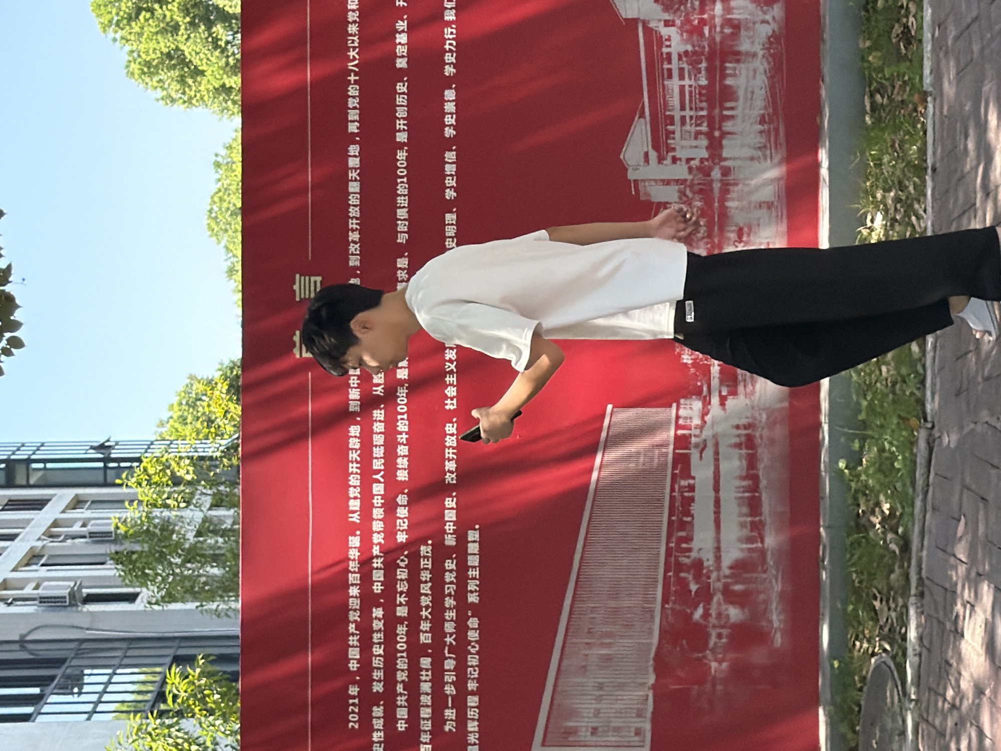 The image shows a young person in a white t-shirt and black pants walking in front of a large red banner. The banner has Chinese text and a stylized image of buildings reflected in water. The text on the banner is a quote or statement related to the history of the Communist Party of China. The background shows a modern building with windows and trees. The ground is paved with bricks and there is grass and fallen leaves. The person is holding a smartphone in their right hand. The scene appears to be outdoors, possibly in a public area or park. The banner indicates that the event or location is related to a celebration or commemoration of the Communist Party of China's centenary.