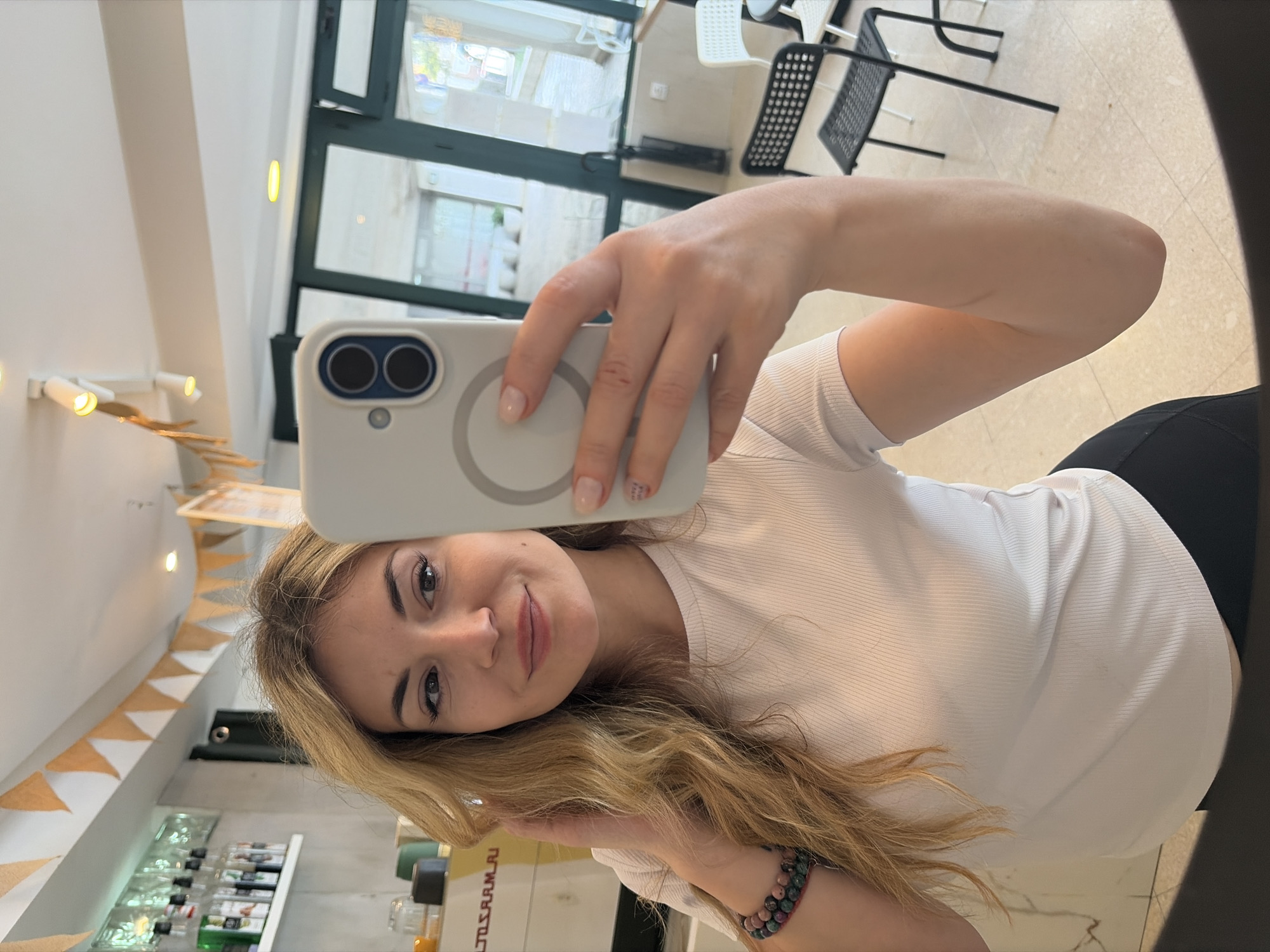 A woman is taking a selfie with a white smartphone. She has long blonde hair, a white t-shirt, and a dark bracelet on her wrist. She is standing indoors, possibly in a cafe or shop in Budva, Montenegro. In the background, there's a window showing a view of the street and chairs. There are also shelves with bottles and decorative flags inside the establishment.