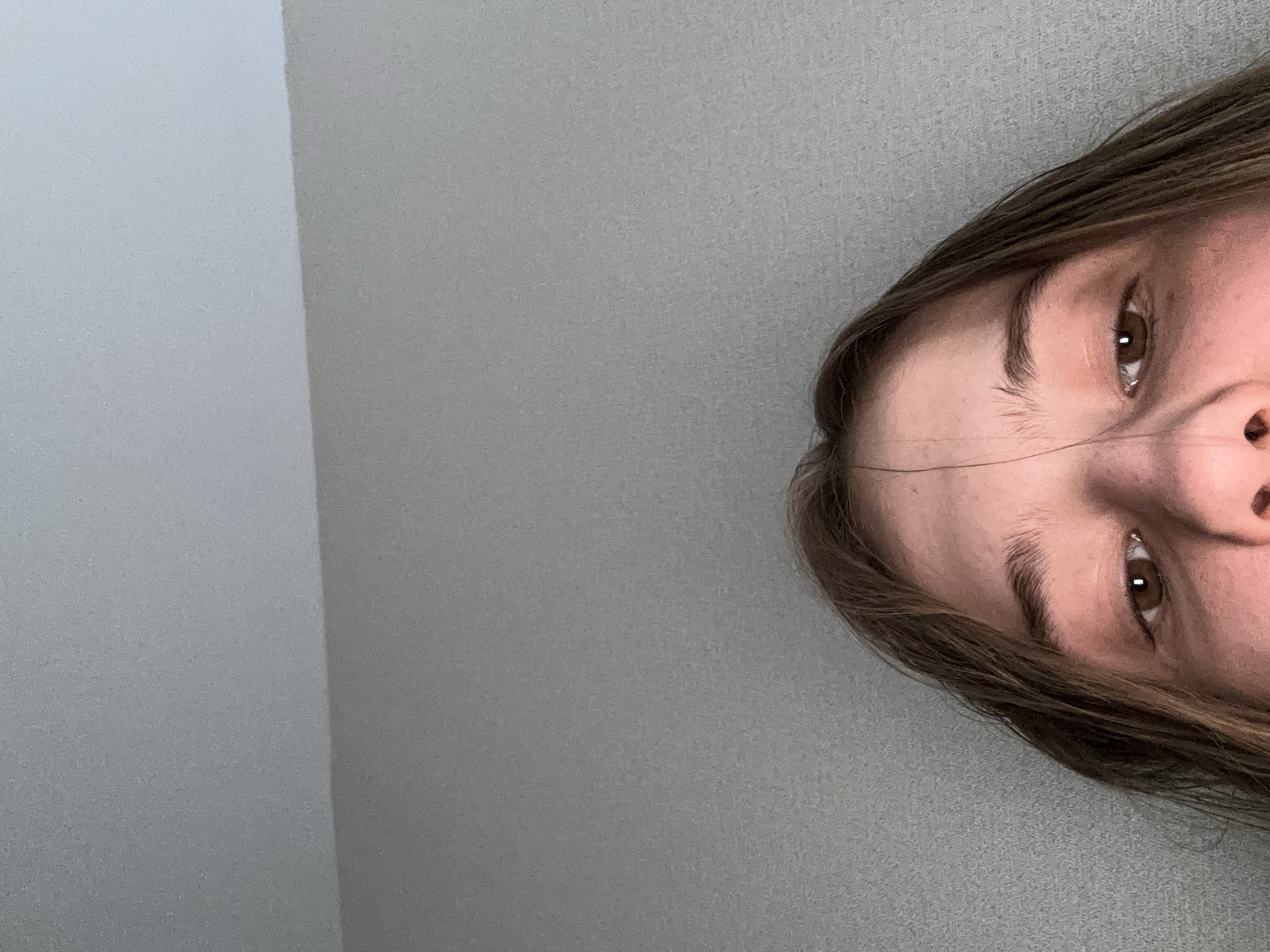 The image shows a close-up view of a person's forehead, eyes, and the bridge of their nose. The person has light brown hair and brown eyes. The background is a textured gray wall, with a slightly lighter gray ceiling meeting it at an angle in the upper part of the image. The lighting is dim and appears to be coming from above and slightly to the left. The image is cropped from the top, obscuring the rest of the person's face and body, as well as any environmental details beyond the wall and ceiling. The image is not located in any specific city.