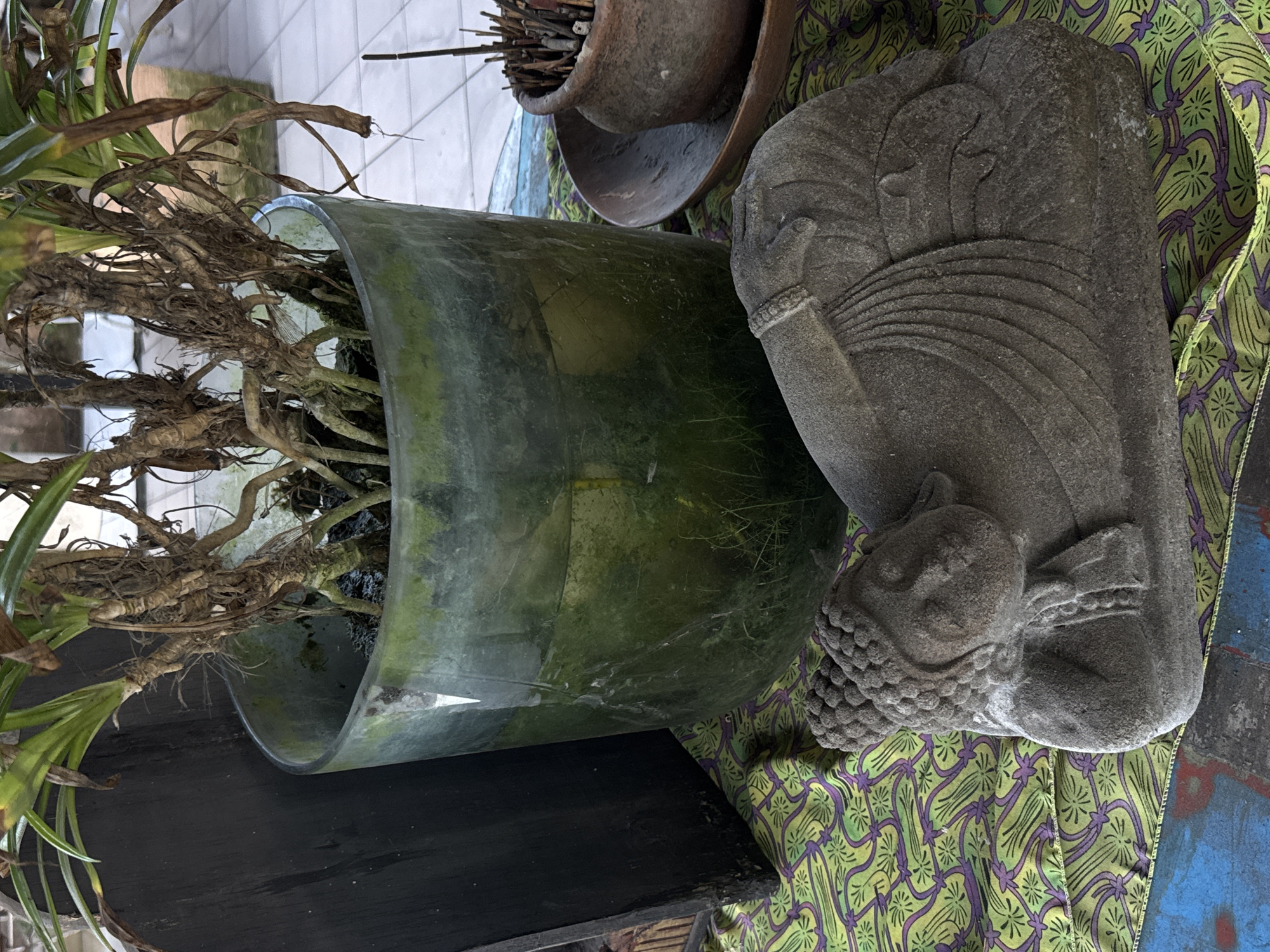The image shows a collection of objects arranged together. A glass vase contains a plant with exposed roots and some green leaves. Next to it is a stone statue of a reclining figure, draped with a patterned fabric in shades of green, yellow, and purple. A small, rough clay pot filled with sticks is in the background. The setting is likely in Kuta Selatan, Indonesia, as this information was provided. The objects are placed on a dark-colored surface. The overall lighting suggests an indoor or sheltered environment.