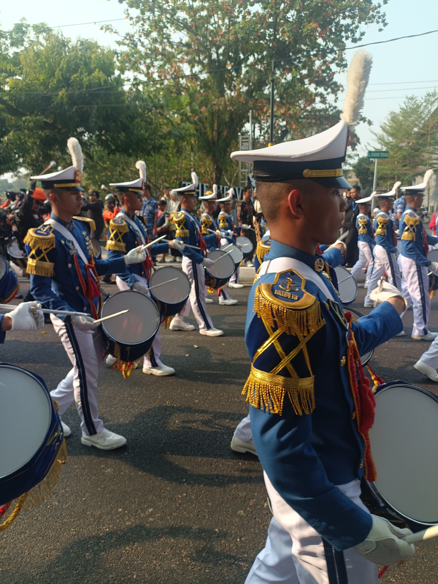 This image depicts a marching band parade in Sleman, Indonesia. The foreground features several young men in blue and white uniforms with gold trim, wearing white hats adorned with large white feathers. They are holding drumsticks and appear to be playing large bass drums slung around their necks. The uniforms are elaborate, with epaulets and decorative tassels on the shoulders and chests. The band members are walking on an asphalt road, with trees and foliage visible in the background. Other people, some on motorcycles and others on foot, are seen lining the street, suggesting a public event. The lighting suggests it is daytime, likely late morning or afternoon, with sunlight casting shadows on the road. The overall scene captures a moment of a vibrant parade.