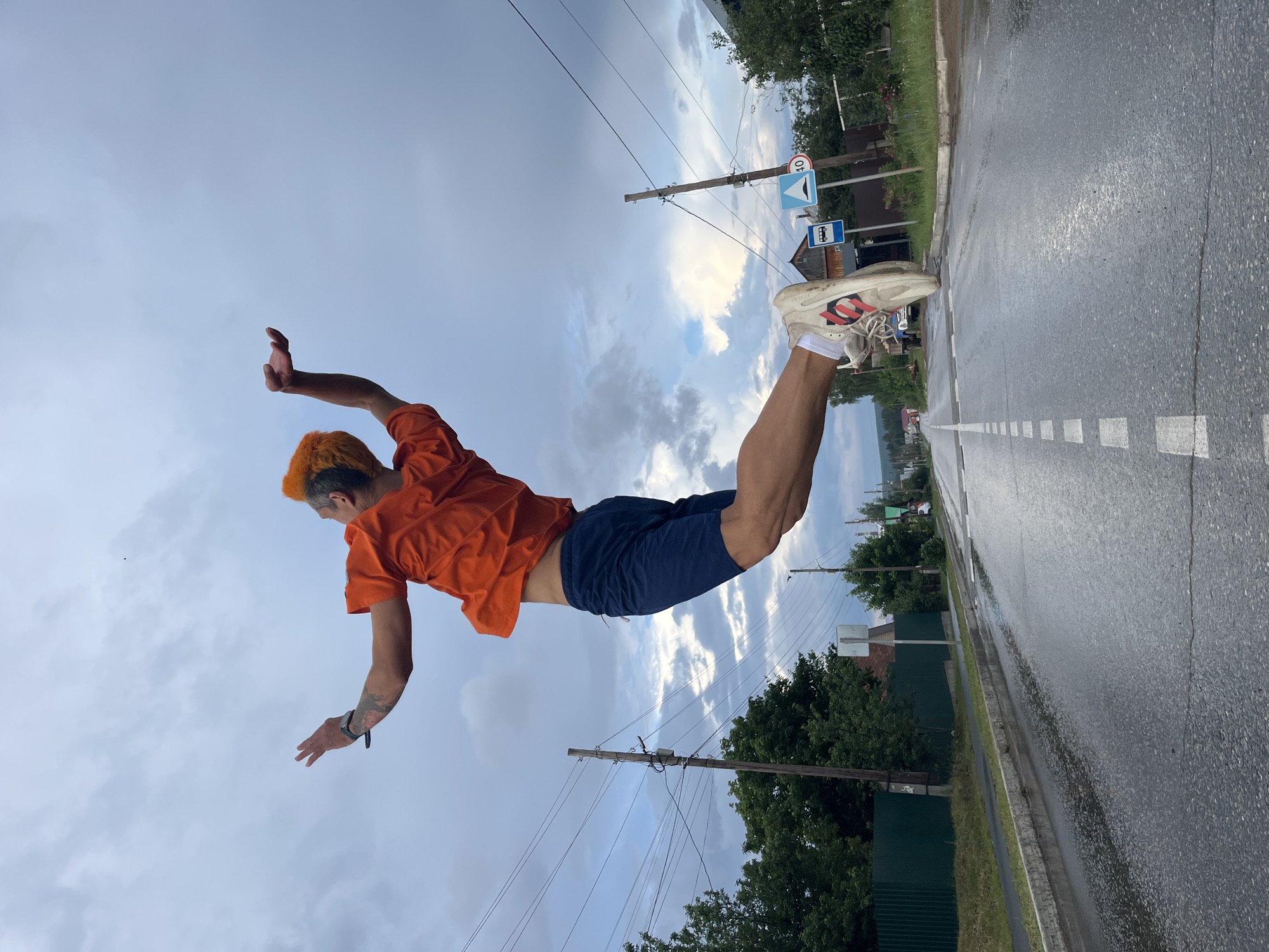A person with bright orange hair is captured mid-air, mid-jump, over a wet, asphalt road. They are wearing an orange t-shirt and dark blue shorts. Their arms are outstretched, and their legs are bent. The background shows a cloudy sky, trees, utility poles with power lines, and a residential area with houses. Traffic signs, including one indicating a speed limit of 40, are visible on the side of the road. The scene appears to be in a rural or semi-urban area, possibly on the outskirts of the city of Chaikovsky, Russia, given the architectural style of the houses and the type of vegetation. The road surface is reflective due to recent rain.