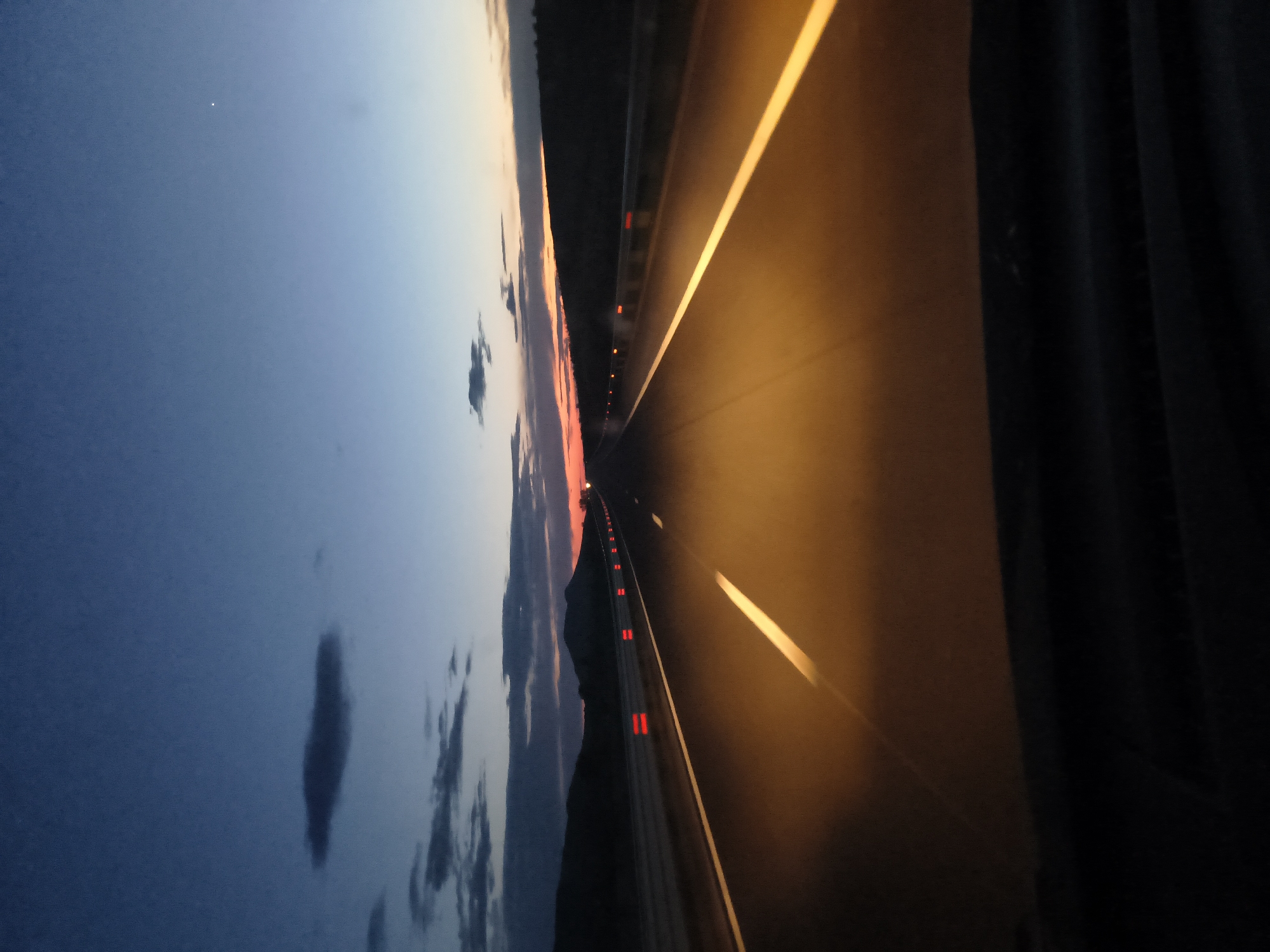 The image depicts an empty highway at twilight, presumably near Gradishte, Bulgaria, though specific city features are not visible. The sky transitions from a deep blue to an orange-red near the horizon, with scattered clouds. The highway, illuminated by the ambient light, has a yellow center line and side lines. Red reflective markers are visible on the guardrail along the highway, indicating the road's edge. The view is from a vehicle or roadside location, positioned at an angle relative to the road.