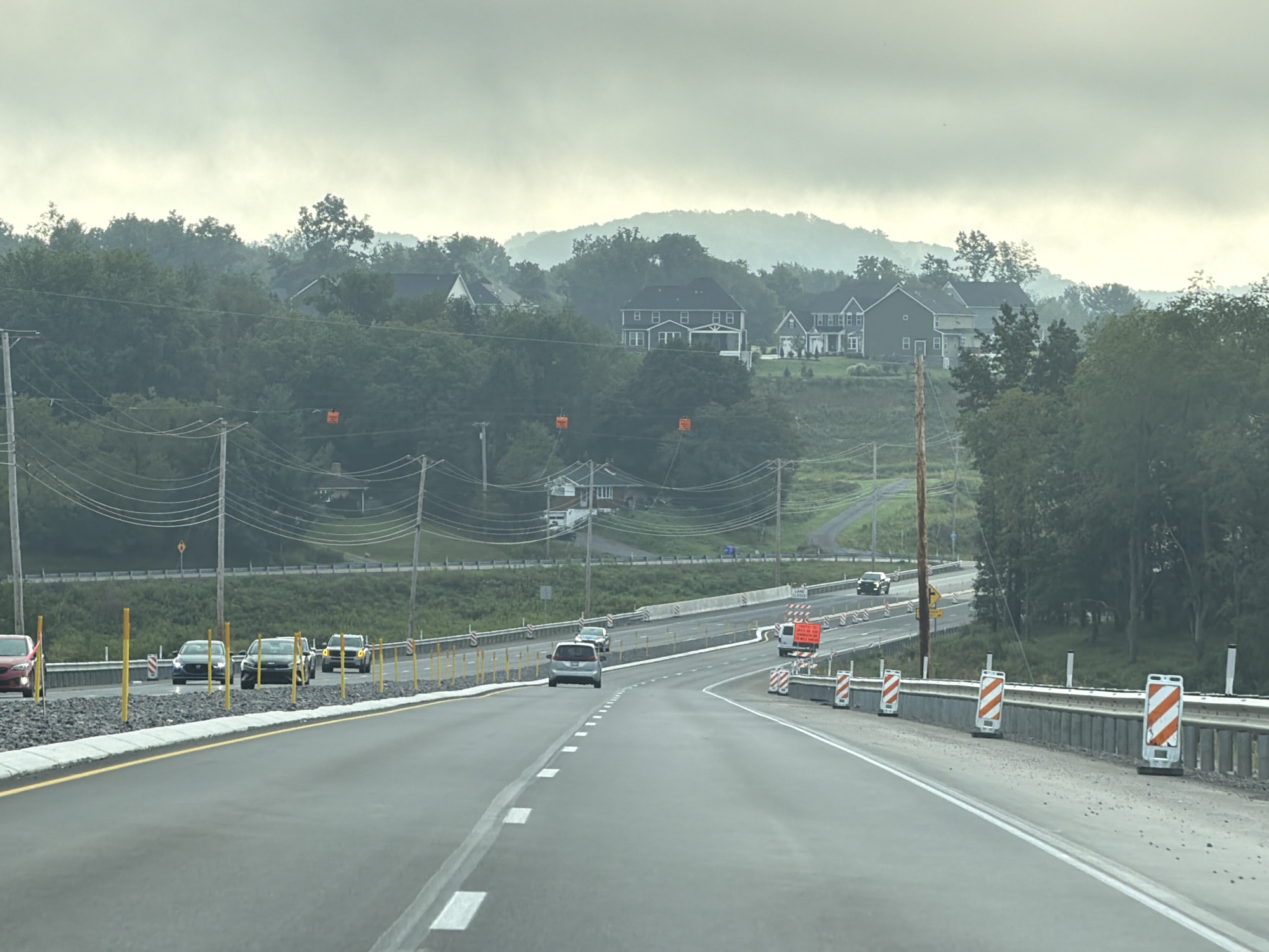 The image shows a view from a vehicle traveling on a highway under construction. The highway has multiple lanes and is bordered by a concrete barrier with orange and white striped delineators. Construction barrels and signs are visible along the roadside. In the distance, there are trees, houses, and rolling hills under a hazy sky. Several cars are on the highway, some of which appear to be moving. Utility poles and power lines are also present. The scene is overcast, suggesting a cloudy day. The location is identified as Middlesex Township, United States of America.