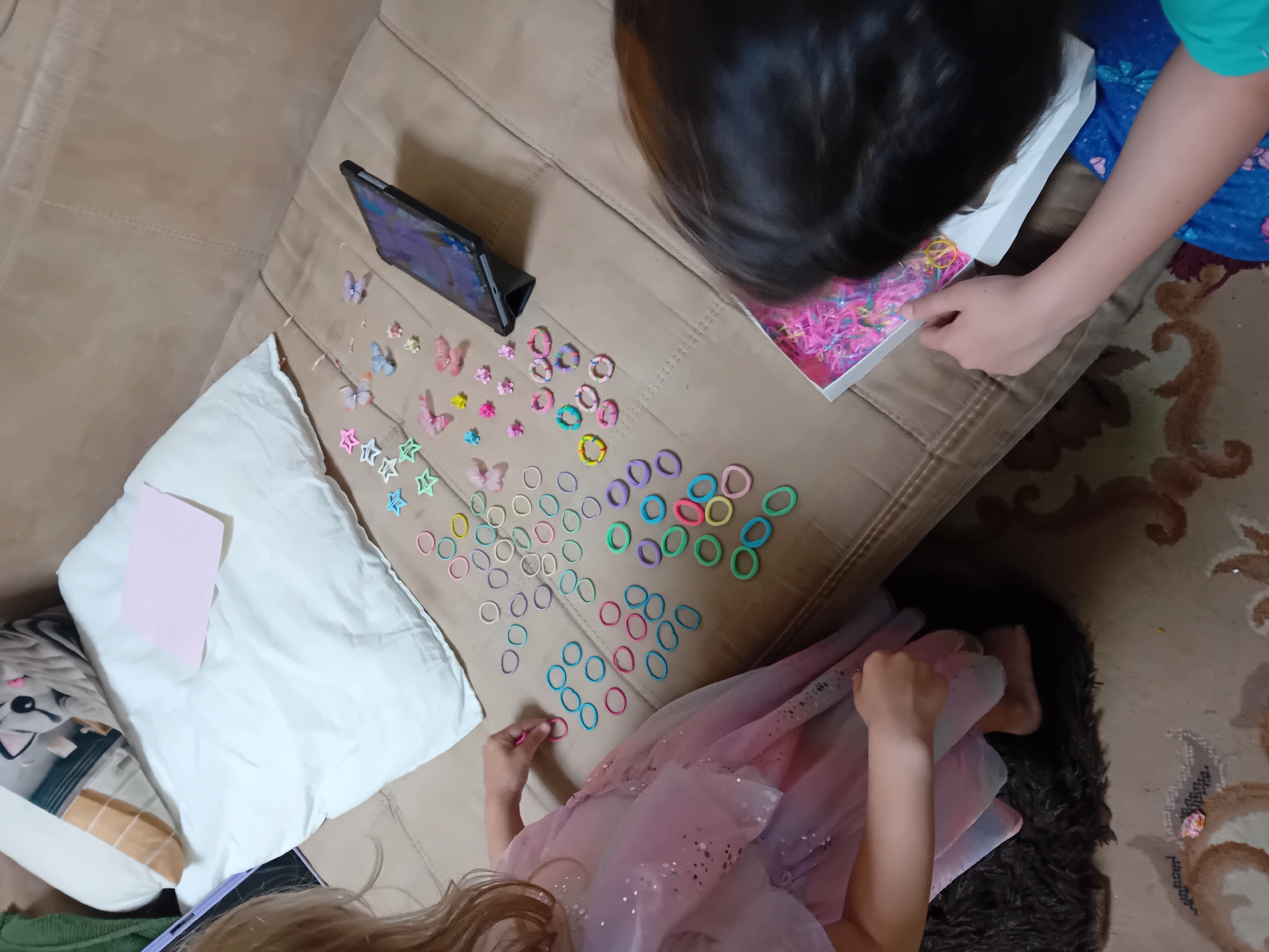 The image shows two young children, likely girls, on a beige upholstered couch. The child on the left, with blonde hair, is wearing a pink sparkly dress and is reaching for colorful rubber bands scattered on the couch. The child on the right, with dark hair, is leaning over a white box filled with colorful rubber bands and appears to be interacting with them. A tablet on a stand is visible on the couch between the children. The background includes a white pillow with a patterned cushion on it and a corner of a patterned rug on the floor. The scene appears to be indoors, likely in a home environment. There is no indication of the city of Chuhuiv, Ukraine, in the image itself.