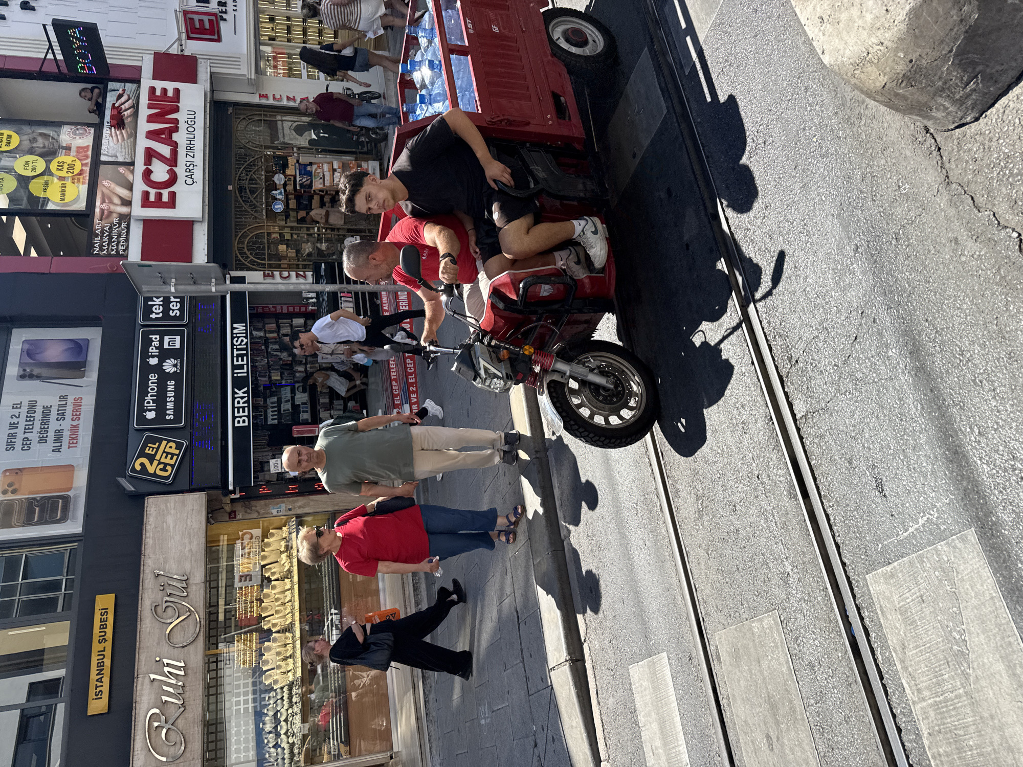 The image captures a street scene in Kadıköy, Turkey, likely in the commercial district given the prominent storefronts. In the foreground, a red three-wheeled motorized vehicle is positioned on tram tracks, carrying several large blue water bottles in its cargo bed. Two men are seated on the vehicle; one is driving, wearing a red t-shirt, and the other, in a black t-shirt, is a passenger. The street appears to be a busy thoroughfare with various shops lining the sides. One shop prominently displays "Ruhi Gül" with a collection of jewelry in its window. Another store has signage for "ECZANE" (pharmacy). Pedestrians are visible in the background, walking along the sidewalk and in front of the shops. The day is sunny, casting distinct shadows on the street and pavement. The architecture suggests a Mediterranean or Middle Eastern urban environment.