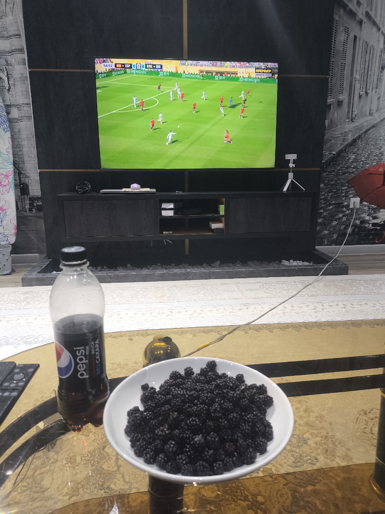 The image depicts an indoor scene, likely a living room or entertainment area, with a large television screen displaying a soccer match. The television is mounted above a dark entertainment unit. In the foreground, a glass table holds a bottle of Pepsi and a white bowl filled with blackberries. A remote control is also visible on the table. The overall ambiance suggests a relaxed setting for watching sports. There is no visible indication of the city of Dushanbe, Tajikistan.