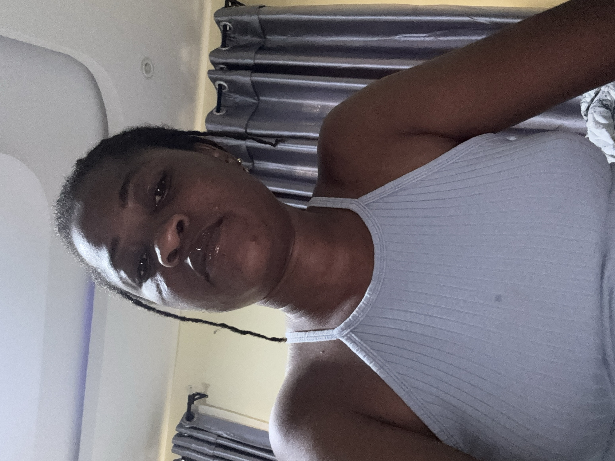 The image is a close-up, low-angle selfie of a woman indoors. She has dark skin and her hair is braided. She is wearing a light blue ribbed tank top. Her lips appear to be glossed. The background shows a ceiling with indirect lighting, and behind her are grey curtains. There is a faint stain on the front of her tank top. The lighting suggests it is daytime, but the indoor setting makes it impossible to determine the exact time. The overall environment suggests a residential bedroom.