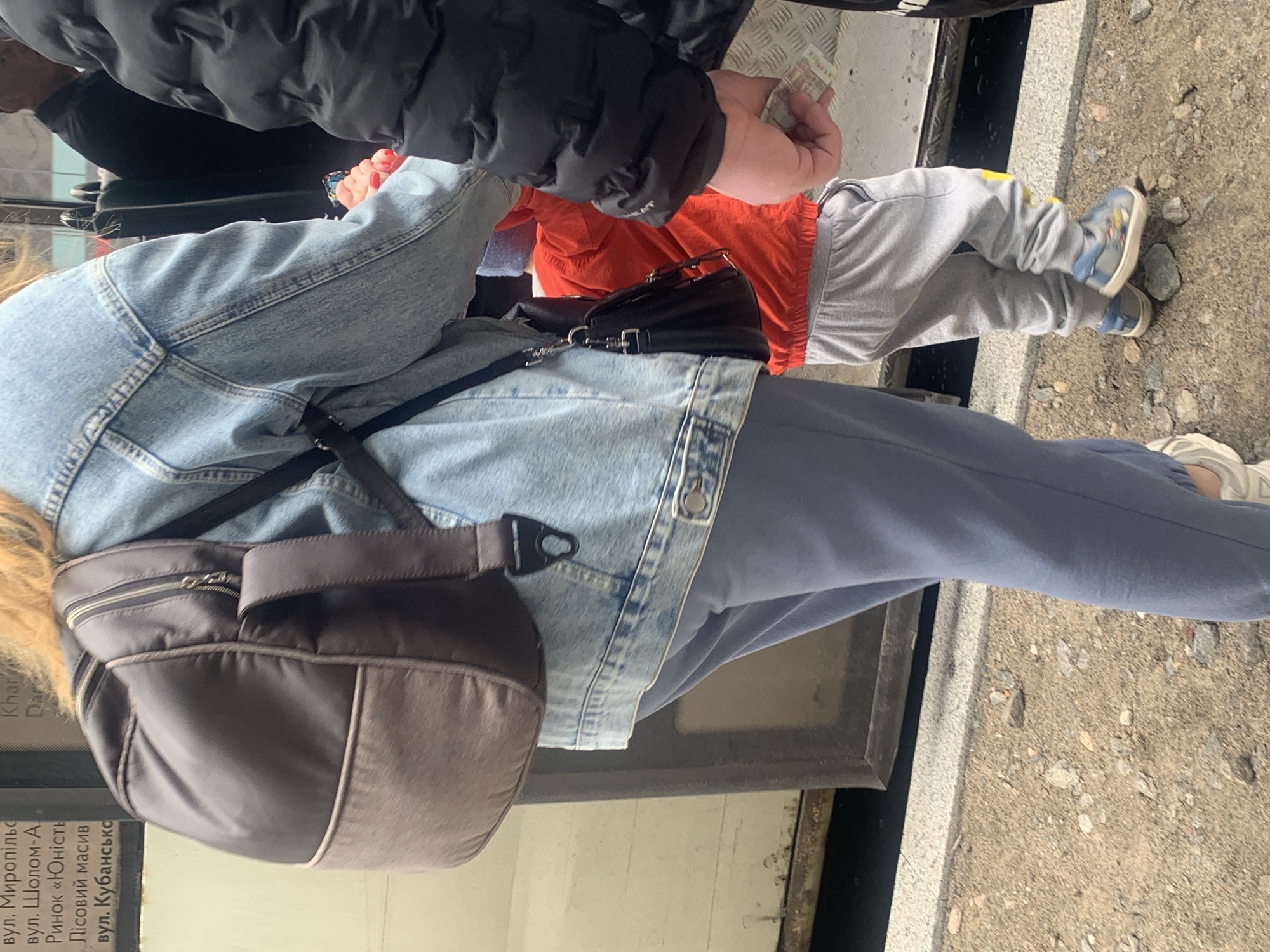 The image shows a person paying with Ukrainian currency, possibly for a bus ticket in Kyiv, Ukraine.
The scene appears to be inside a public transport vehicle, possibly a bus or tram, in Kyiv, Ukraine. The person is handing Ukrainian money to someone, presumably a driver or conductor, likely to pay for the fare. A child in gray sweatpants and an orange shirt is also present. The person paying is wearing a light blue denim jacket and a dark blue tracksuit bottoms, with a dark-colored handbag or rucksack. The bottom left corner of the image shows a partial view of a sticker or sign with Ukrainian text, including street names: вул. Миропільс (street Myropils), вул. Шолом-А (street Sholem-A), Ринок «Юність (Yunist Market), Лісовий масив (Forest Massif), вул. Кубансько (street Kubansko).