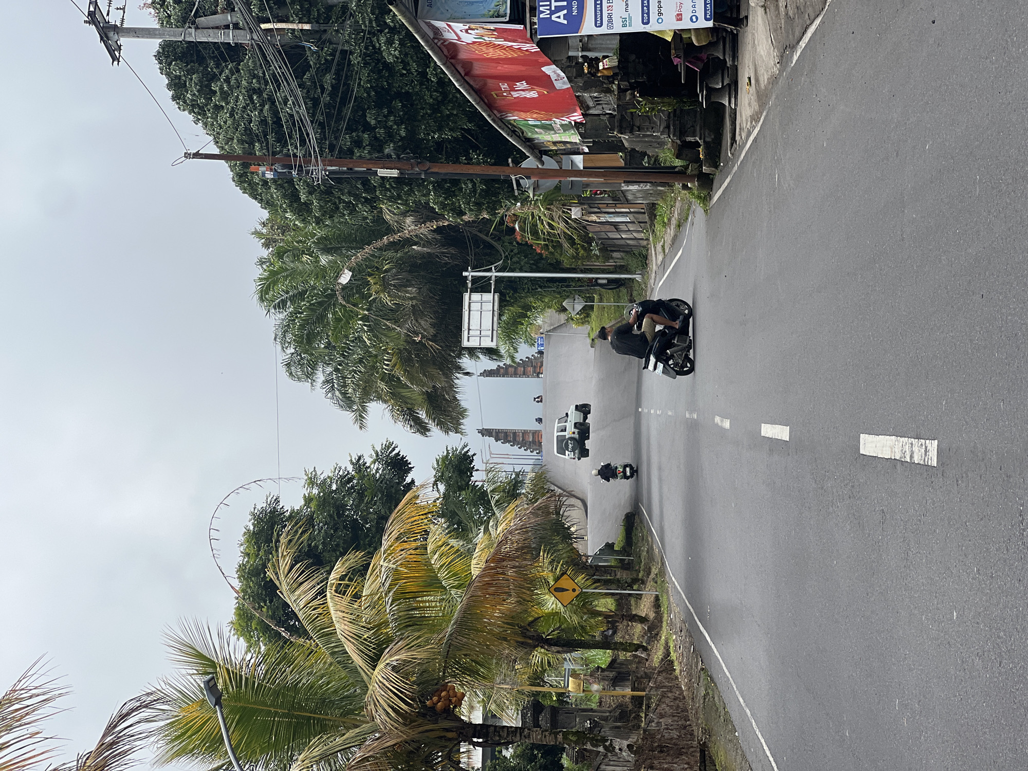 A paved road curves uphill in Rendang, Indonesia, under a gray, overcast sky. Palm trees line the left side of the road, some bearing coconuts. A yellow diamond-shaped sign with a black exclamation mark is visible near the trees. On the right side of the road, shops with red banners and signs are situated.
Several vehicles are present on the road. In the foreground, a person rides a black scooter, moving uphill. Further up the road, another scooter is visible, with two people on it, also heading uphill. A white SUV is also seen ascending the hill. At the crest of the hill, an ornate gateway structure is visible. Electrical poles and wires are present on the right side of the road. The scene depicts a typical street view in the area.