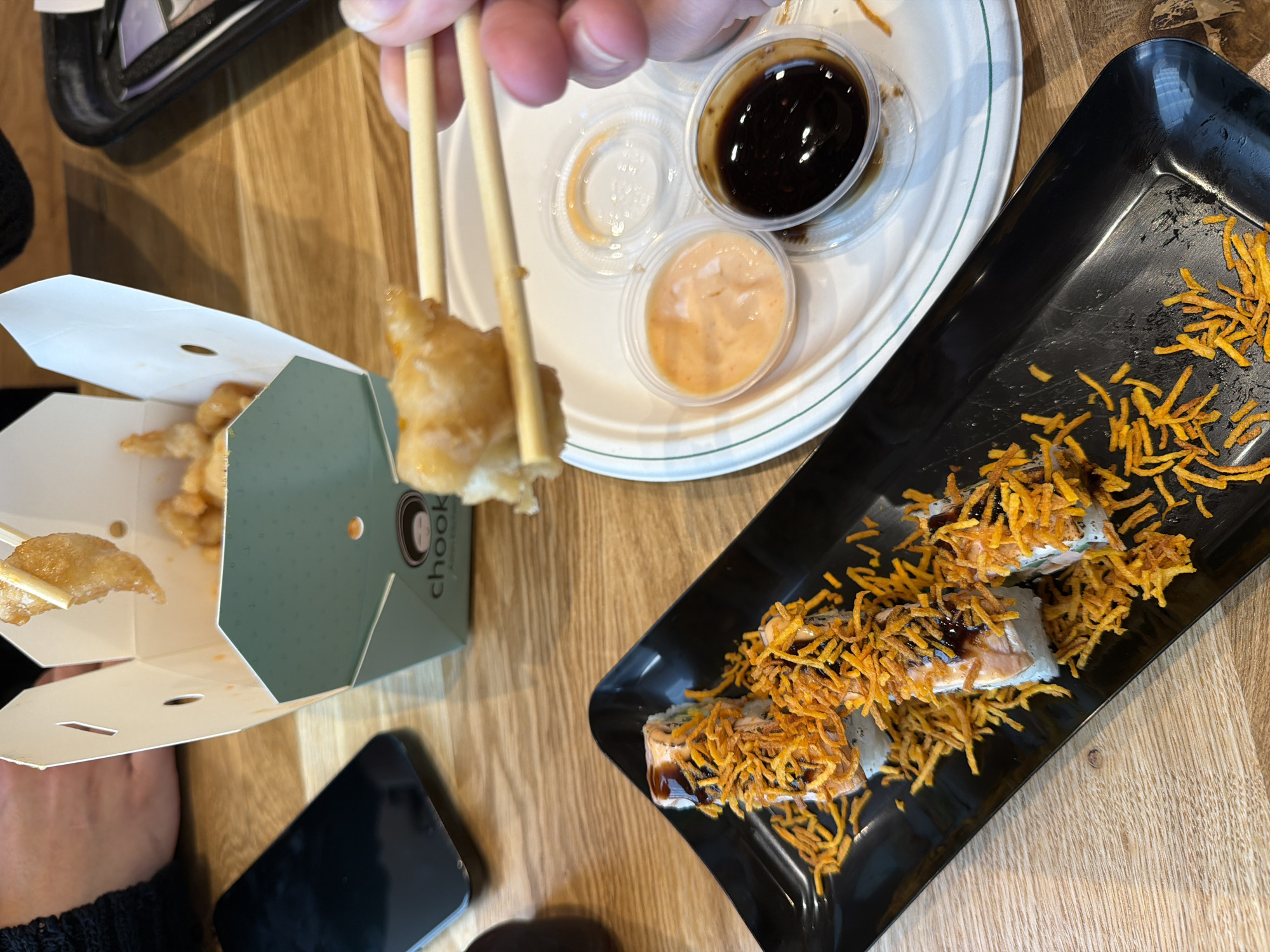 The image depicts a table setting, likely in a restaurant, featuring a partially eaten order of "Chook" brand fried chicken in a takeout box, a plate of sushi topped with fried noodle-like strands, and a sauce plate with three small containers holding dipping sauces (soy sauce, a light orange sauce, and an empty container). A person is using chopsticks to pick up a piece of the fried chicken. A black smartphone is visible near the takeout box. The restaurant is located in Tel-Aviv, Israel, based on the "Chook" branded takeout box. The table has a light wood grain finish.
