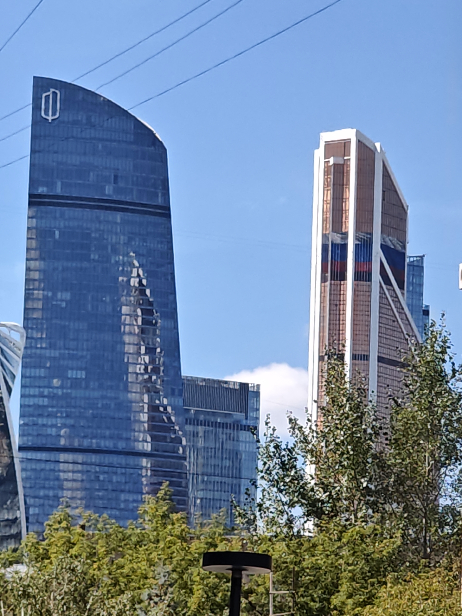 The image captures a sunny day in Moscow, Russia, showcasing the modern architecture of the Moscow International Business Center, also known as Moscow City. In the foreground, lush green trees with vibrant foliage partially obscure the lower portions of several skyscrapers.
Dominating the left side of the frame is a tall, dark blue glass skyscraper with a distinctive curved silhouette. A white, stylized logo, resembling an open tulip or a shield with a central vertical line and two flanking curves, is prominently displayed near the apex of this building. The building's facade reflects the sky and the surrounding structures, creating a dynamic visual effect.
To the right of the blue skyscraper stands another impressive tower with a more angular design. Its exterior features a combination of copper-toned panels and white accents, with vertical stripes of red, white, and blue, reminiscent of the Russian flag, integrated into its design.
Further back, other modern skyscrapers of varying heights and architectural styles can be seen, contributing to the impressive urban skyline. The sky is a clear, bright blue, with a few wispy clouds adding texture. Overhead, several parallel power lines stretch across the upper part of the image, a common sight in urban environments. The overall impression is one of a contemporary and bustling cityscape.