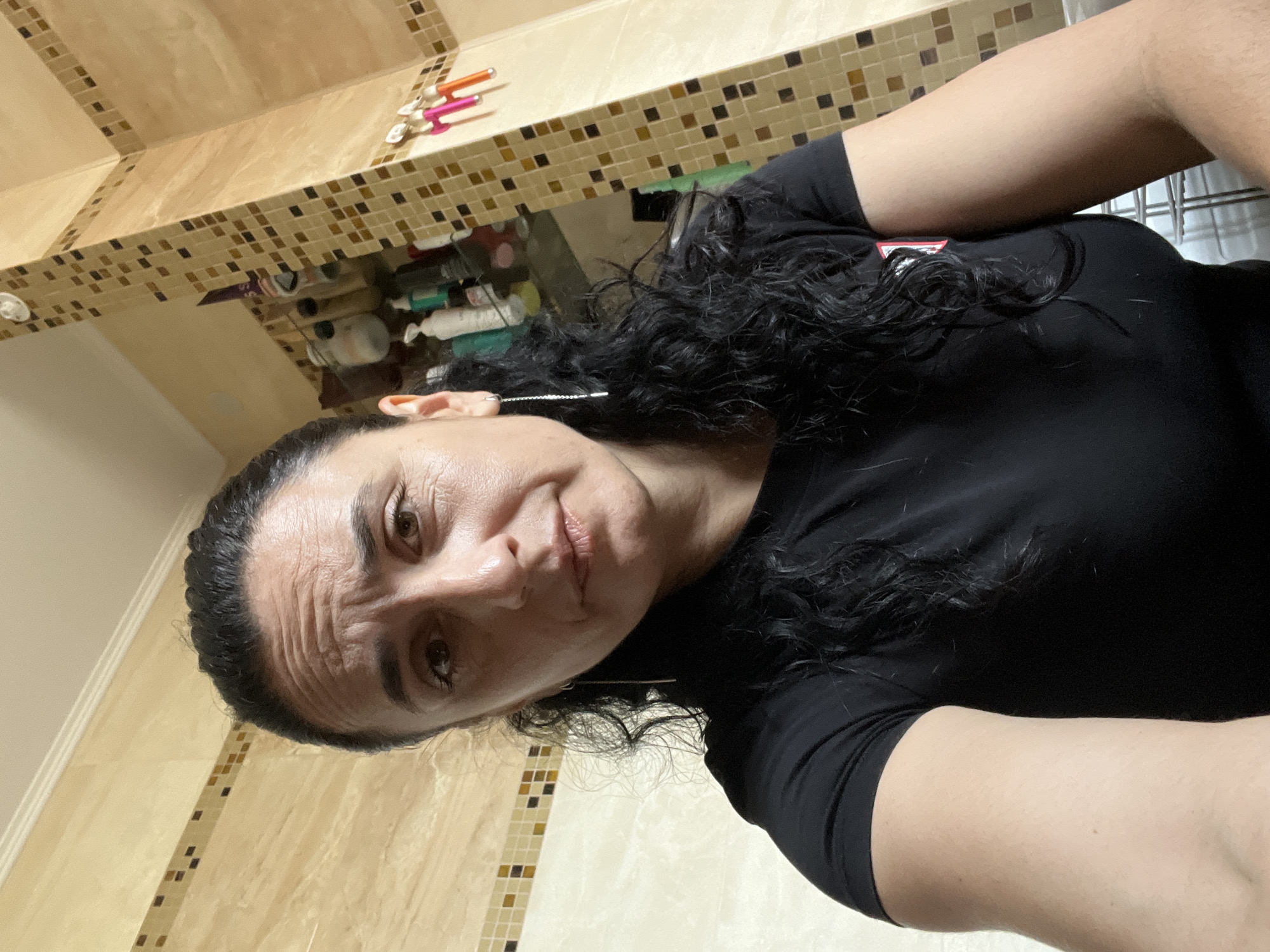 A medium shot depicts a woman with dark, curly hair, wearing a black t-shirt, presumably in a bathroom. The environment is distinguished by beige and brown mosaic tile walls. Hair styling tools, and assorted cosmetic bottles are visible on a shelf in the background. The image does not provide enough information to determine the location in Onokivtsi, Ukraine.