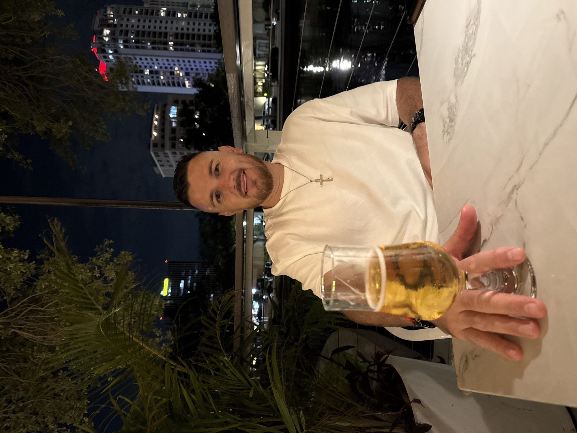 A nighttime photograph depicts an adult male seated at an outdoor table in Saint Petersburg, Florida, United States of America. The subject, a man with short dark hair, a trimmed beard, and fair complexion, is wearing a plain white short-sleeved t-shirt, a silver-toned crucifix pendant on a chain, and a dark-colored wristband on his left arm. He is smiling directly towards the camera. His right hand rests on the stem of a clear glass containing a pale amber, carbonated beverage, likely beer, with a thin layer of foam at the top. The table surface is light-colored, possibly white marble or engineered stone with grey veining.
To the left and behind the subject, lush green foliage, including palm fronds, is visible. In the background, the illuminated city skyline of Saint Petersburg is discernible against a dark sky. Prominent features include a tall, white high-rise building with multiple lit windows and distinctive red lighting at its apex, and another building with a yellow illuminated sign near its top. To the right of the subject, a dark, reflective surface, consistent with water, mirrors the city lights. A dark metal railing runs horizontally across the mid-ground, separating the table area from the distant view. Vehicle lights are visible on a street or roadway in the distant background.