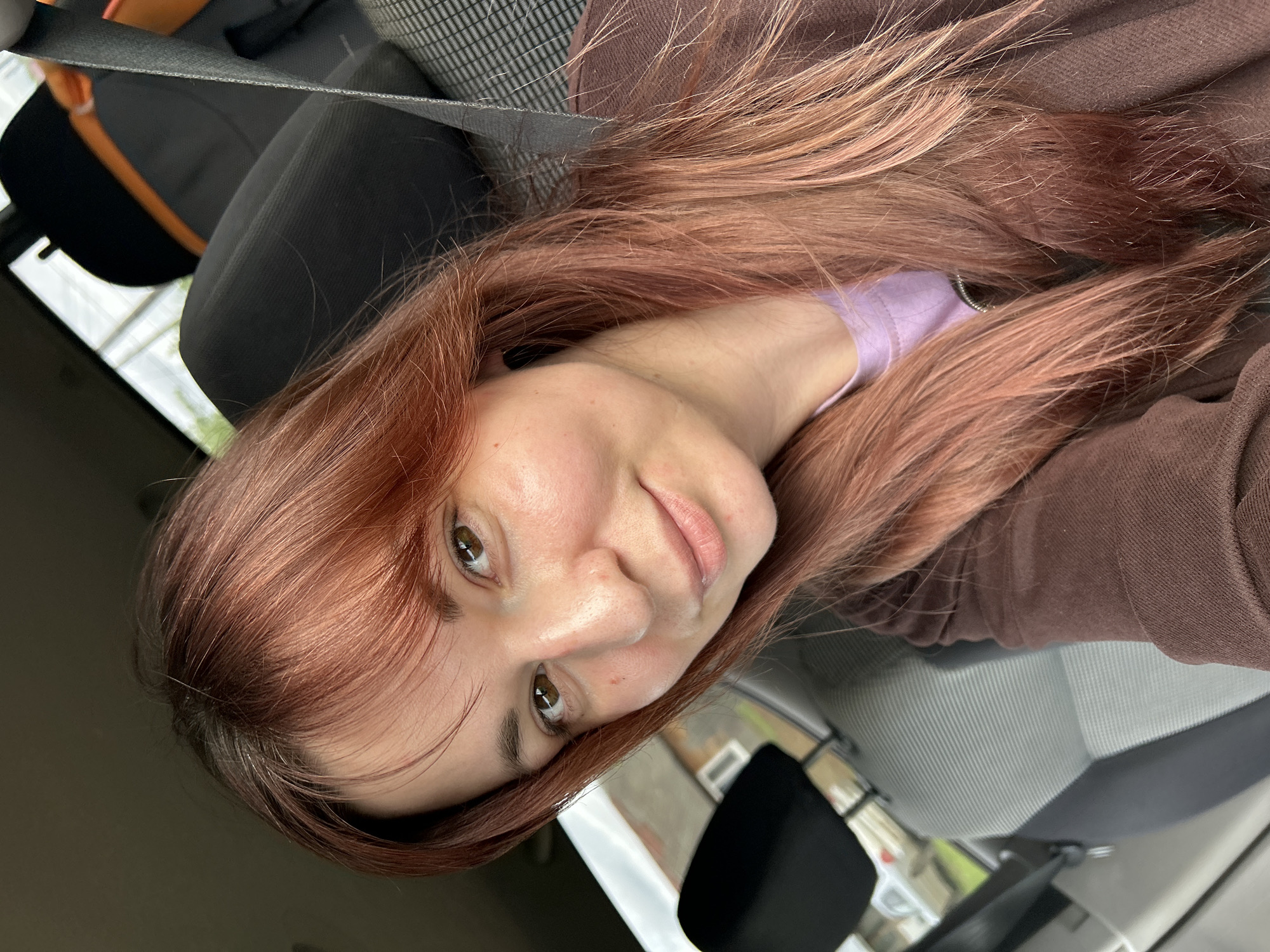 A female individual with long, reddish-auburn hair, styled with bangs, is depicted in a self-portrait within the passenger compartment of a vehicle. Her eyes are brown, and she has a light complexion, looking directly at the camera with a subtle, closed-mouth smile. She is wearing a brown long-sleeved outer garment over a light purple top. The car interior features gray and white striped fabric seating, black headrests, and a dark seatbelt visible across her right shoulder. An orange object is partially visible behind the front passenger seat. Through the car windows, a blurred view of exterior residential or urban buildings and a street is discernible. This scene is located in the city of Фоки, Russia.