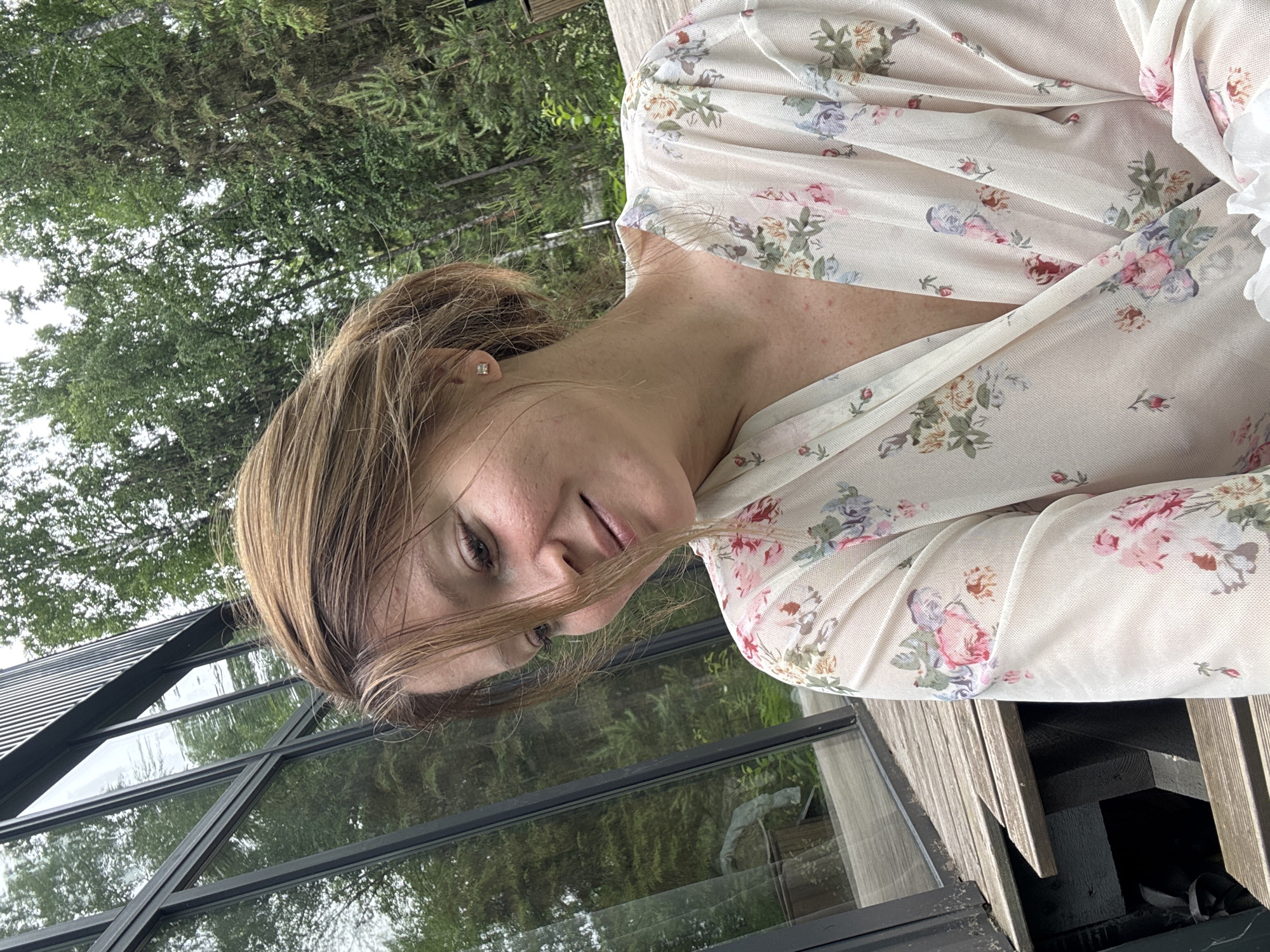 A woman with light brown hair, wearing a light-colored robe with a floral print, smiles and looks down. She is positioned on a wooden deck, adjacent to a modern building with large glass windows. The background consists of lush green trees. The scene is set outdoors, in what appears to be a natural environment, possibly a residential area in Bakhtiyary, Russia.