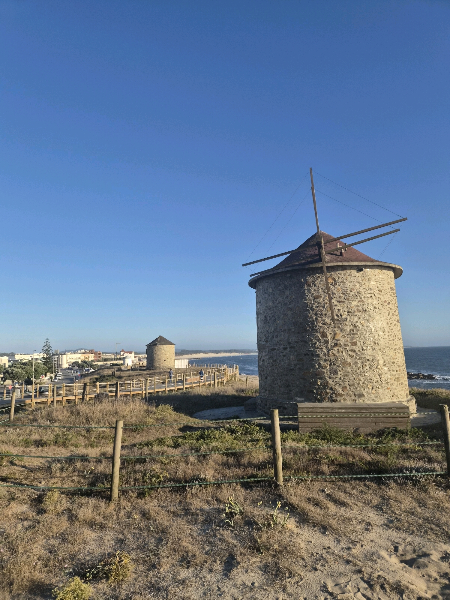 This is a vertical, eye-level, medium shot of two stone windmills with conical roofs located in Esposende, Portugal. The windmill in the foreground is larger and closer to the viewer, while the second windmill is smaller and in the background to the left. Both windmills have visible wooden support beams extending from their roofs, but their sails are absent.
The windmills are situated on a grassy, sandy hill overlooking the ocean. A wooden boardwalk with railings runs parallel to the shoreline in the middle ground, separating the grassy area from the beach. Several people are visible walking on the boardwalk.
In the background, a coastal town with buildings of varying heights and colors is visible along the coastline. The ocean is a calm blue, meeting the horizon under a clear, bright blue sky. The foreground is characterized by dry, sparse grass and sand, with a wooden fence and posts interspersed. The lighting suggests it is daytime, with the sun casting a warm light.