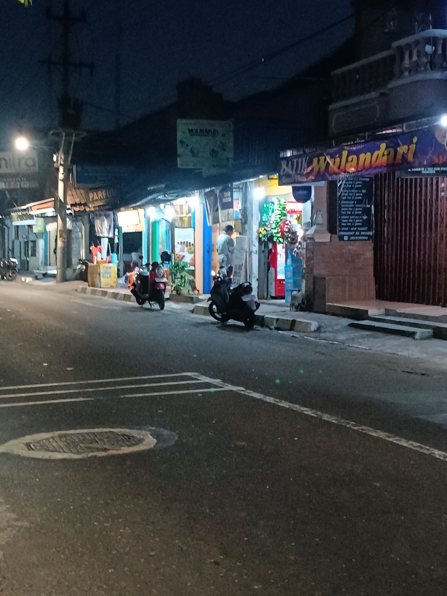 The image depicts a street scene at night in Yogyakarta, Indonesia, likely in a commercial area with shops lining one side of the road. Several motorbikes are parked along the sidewalk, and some are in motion. One motorbike, a red scooter, is stopped on the sidewalk, with a person wearing a red jacket seated on it. Another black scooter is parked on the sidewalk in front of a shop.
The shops display various goods, with one sign reading "BATIK WULANDARI" indicating a batik shop. Another sign visible reads "PARFUM." The shops are illuminated, creating a somewhat festive atmosphere with colorful displays, possibly indicating a market or a shopping street. There are people visible in the shops, though their actions are not clearly discernible due to the low light conditions. The road surface shows painted lane markings and a manhole cover. The overall environment suggests a typical urban street in Indonesia at night.