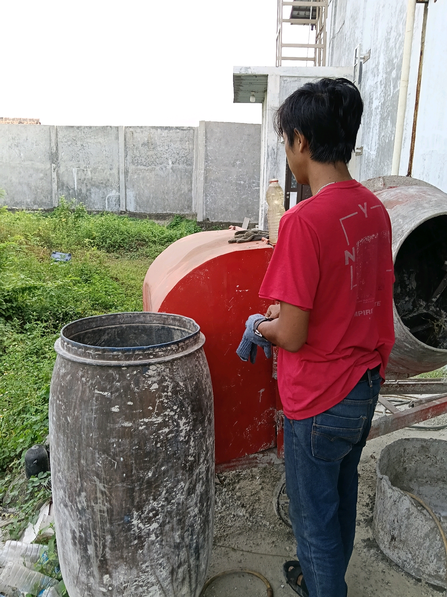 A young male with short black hair, wearing a red t-shirt and blue jeans, stands in an outdoor construction area in Belimbing, Indonesia. He is facing away from the camera, looking towards a cement mixer and a large, dark gray barrel. In his hands, he holds a blue cloth or glove. The environment includes a concrete wall, overgrown green vegetation, a red cement mixer, and a weathered, cylindrical barrel. The ground is a mix of dirt and loose gravel, with some construction materials visible. A white building with a metal staircase is in the background.