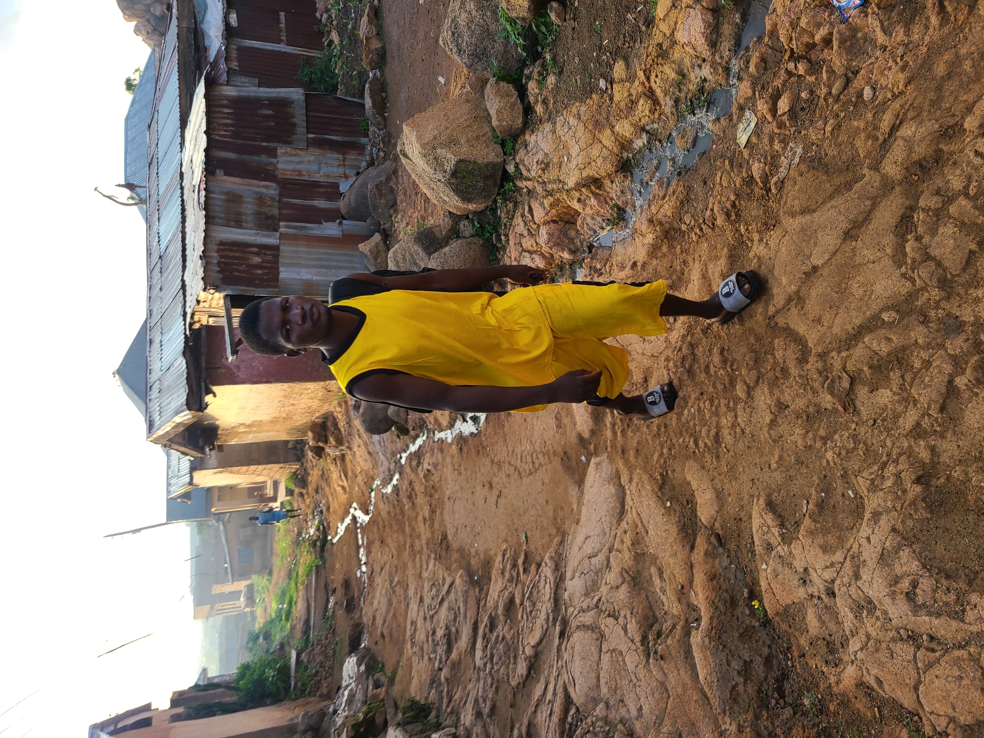 The image depicts a young Black male walking on a dirt path in a residential area of Jos, Nigeria. He is wearing a yellow tank top and yellow shorts, with black and gray slide sandals. The path is rough and uneven, with exposed rocks and a small stream of murky water flowing alongside it. In the background, there are several houses with corrugated iron roofs, some appearing dilapidated. The sky is bright and hazy, suggesting it is either early morning or late afternoon. The overall environment appears to be a low-income urban settlement.