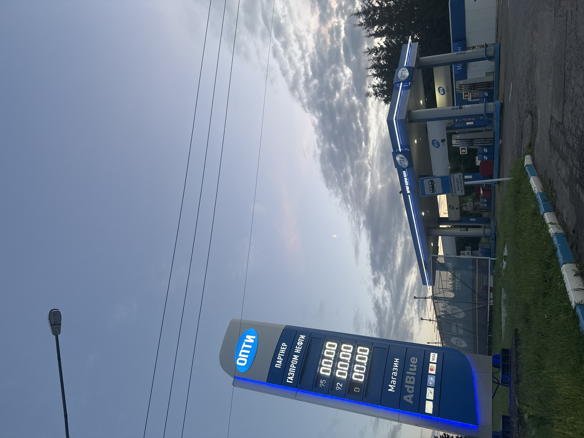 The image shows a gas station in the city of Vereya, Russia. A tall blue and white sign displaying fuel prices and the brand "ОПТИ" (Opti) stands prominently on the left. The sign shows prices for 95, 92, and D (diesel) fuel. Below the fuel prices, the words "Магазин" (Shop) and "AdBlue" are visible. The gas station canopy, also branded with "ОПТИ", is lit with blue lights. Several fuel pumps are visible under the canopy. A sign indicating "ОБЯЗАТЕЛЬНАЯ ВЫСАДКА ПАССАЖИРОВ" (Mandatory Passenger Drop-off) is also present. The sky is overcast with clouds, and a faint crescent moon is visible. There are power lines stretching across the sky. Trees line the background of the gas station. The ground is paved asphalt with some grassy areas.