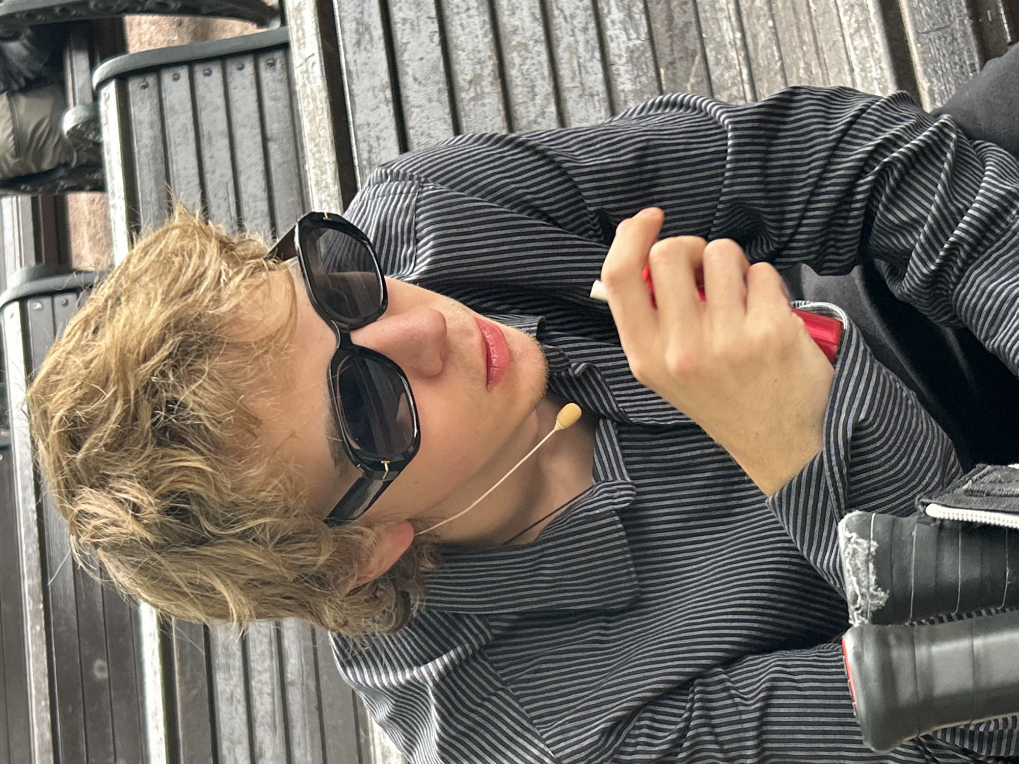 A young person with blonde, messy hair and black sunglasses sits on a wooden bench, wearing a black and gray striped button-down shirt. A thin, light-colored wire with a small, yellow microphone attachment hangs from their ear and drapes down their neck. Their right hand is raised, holding a small, red object. In the foreground, two black objects with ribbed grips, possibly parts of sports equipment like tennis rackets, are visible. The background consists of more wooden benches and a hint of greenery. The scene appears to be outdoors in a public space.