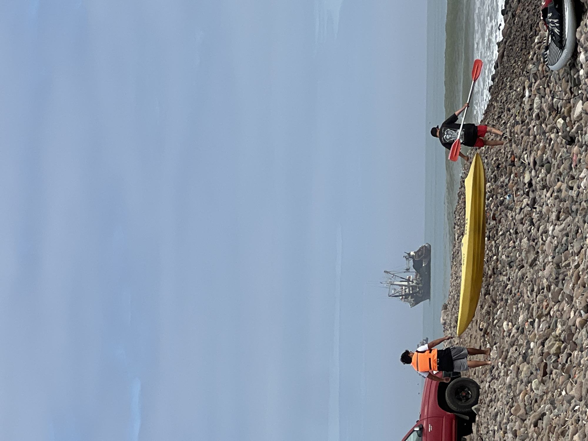 The image depicts a beach scene in Arica, Chile. A yellow kayak lies on a rocky shore. Two people are present; one is wading into the water with a paddle, while the other stands near a red pickup truck. A fishing vessel is visible in the distance. The sky is overcast.