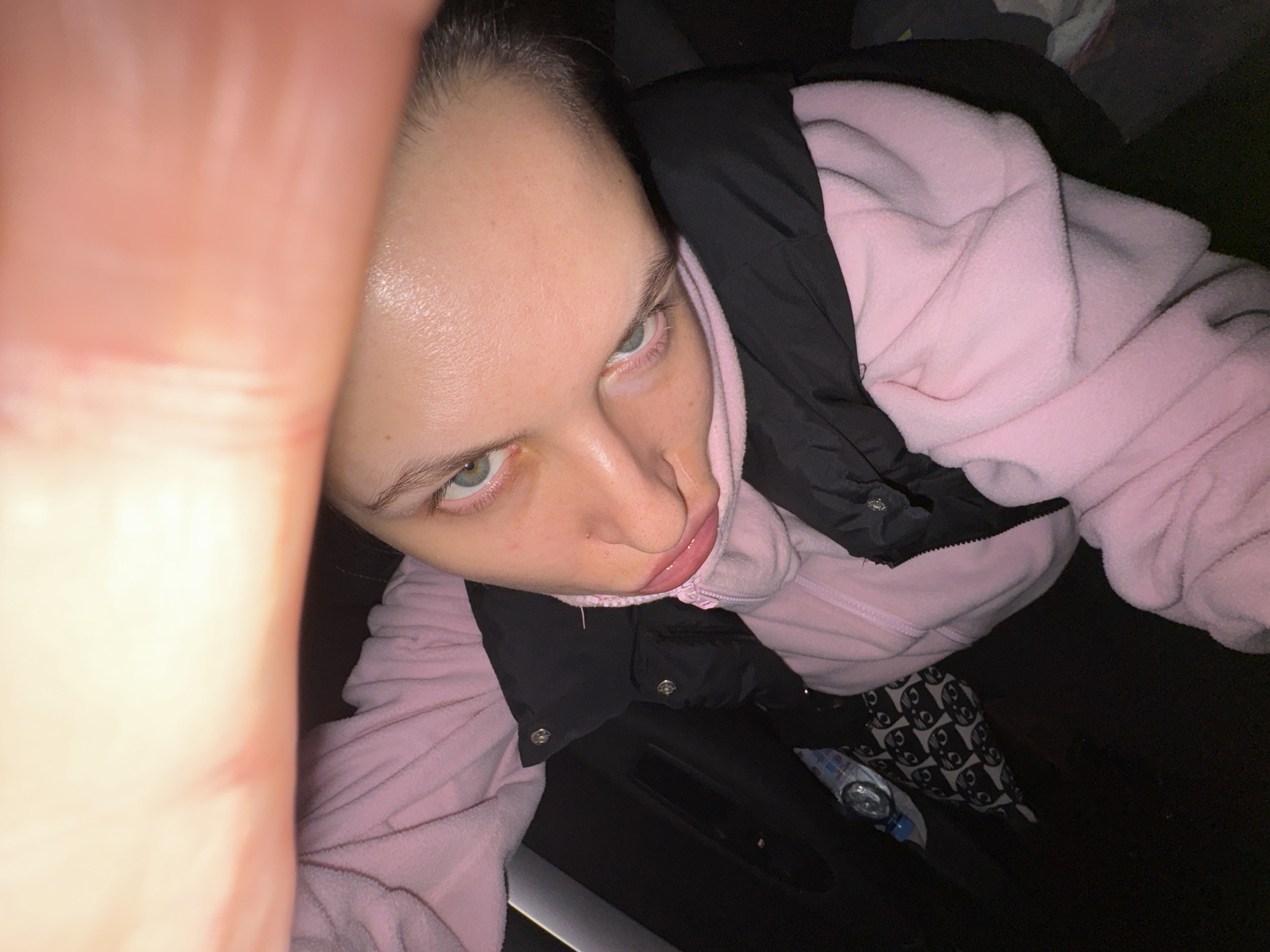 A high-angle selfie depicts a person with light skin, light eyes, and dark hair. They are wearing a pink hooded sweatshirt under a black jacket. Part of a hand or arm is visible in the foreground, partially obscuring the left side of the frame. Below the person, parts of patterned black and white pants and a plastic bottle can be seen, along with a dark surface. The exact location in Uvarovo, Russia cannot be determined from the image.