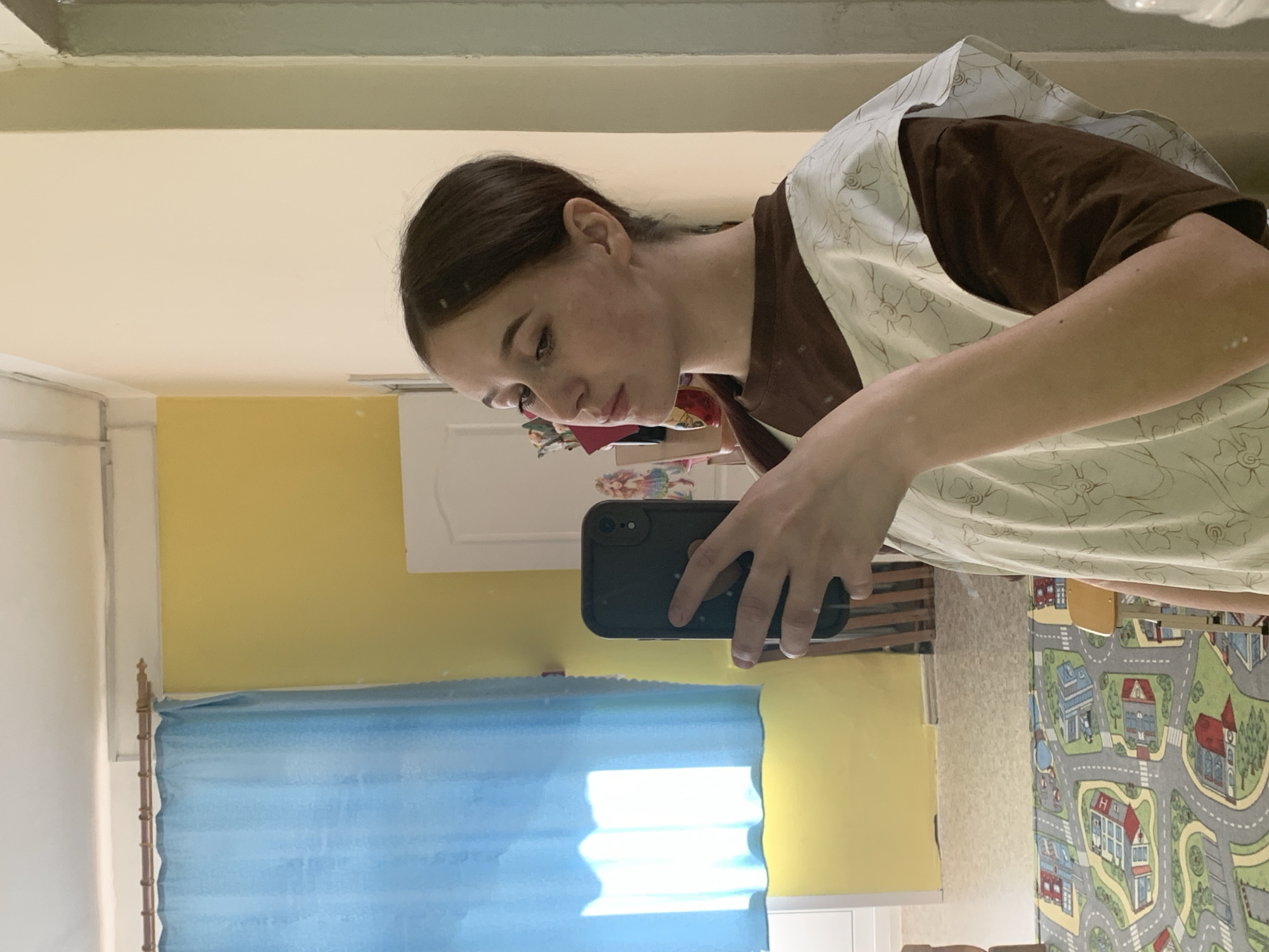 A person is taking a selfie in a room. The person is wearing a brown shirt and a light-colored apron with a floral pattern. The person is looking down at the phone. The room has yellow and white walls, and a blue curtain. There is also a rug with a city map design. The location is Фоки, Russia.