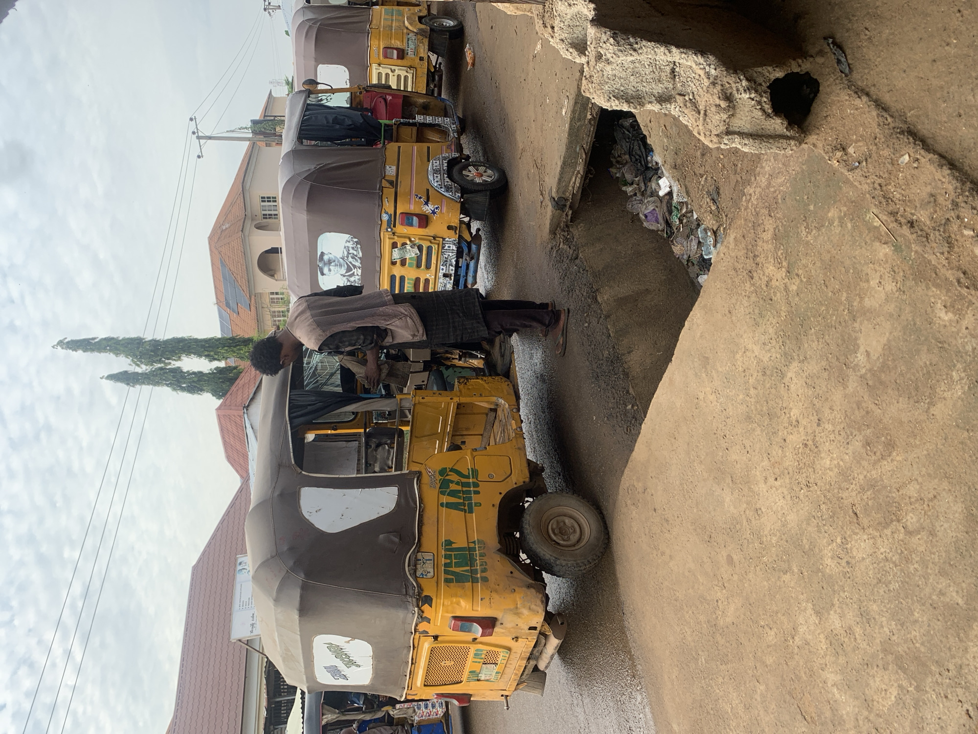 In Abuja, Nigeria, three yellow auto-rickshaws are parked on a wet asphalt road, lined up on the right side of the frame. The lead auto-rickshaw, a KMC 4472 model, is prominently featured in the foreground, facing left. A person, wearing a vest and dark pants, stands to the right of this vehicle, seemingly engaged in an interaction with someone inside. The side of the auto-rickshaw displays a picture of a man in uniform. Behind it, two more auto-rickshaws are visible, also parked. The environment includes buildings with red tiled roofs in the background, power lines overhead, and two tall trees. A drainage ditch filled with trash runs along the right side of the road, adjacent to a concrete structure. The scene appears to be a street in a commercial or residential area, characterized by its modest infrastructure and the presence of common modes of local transportation.