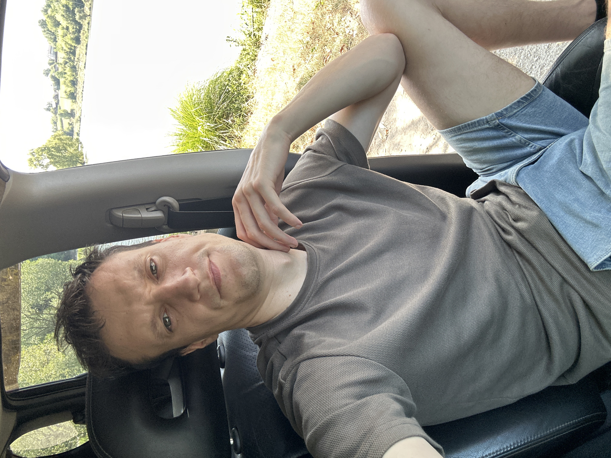 A man is sitting in the driver's seat of a vehicle, taking a selfie. He is wearing a grey t-shirt and denim shorts. His left arm is resting on his knee, and his right hand is touching his chin. The car's interior is visible, including the black leather seat and a grey seatbelt. Outside the car window, a body of water is visible, with trees and some buildings in the background. The location is Makiv, Ukraine, as indicated by the provided context. The scene appears to be set on a sunny day, with natural light illuminating the environment.
