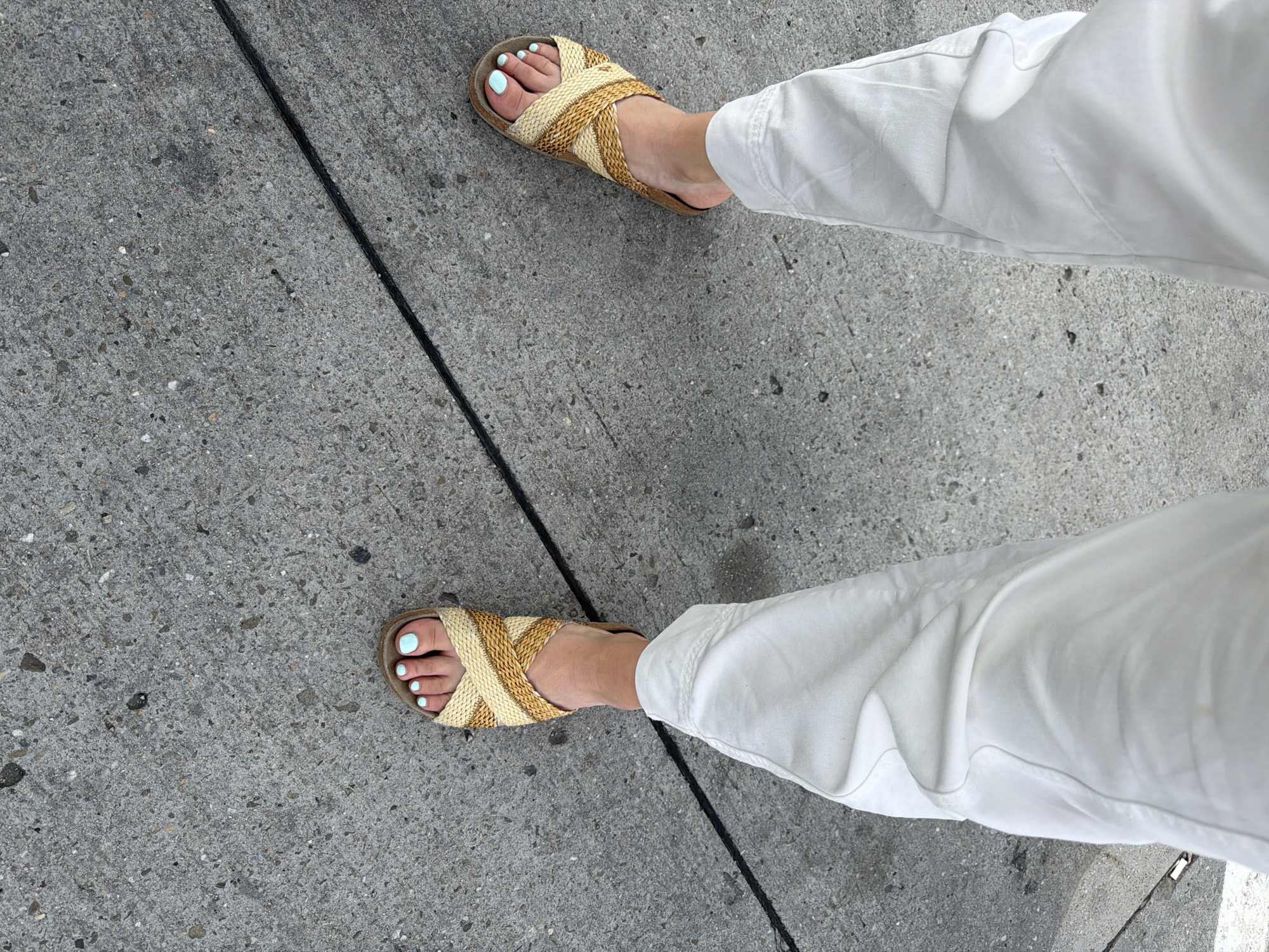This is a top-down, eye-level shot of a person's legs and feet from the knees down, standing on a textured concrete surface. The person is wearing white pants and woven slide sandals. The sandals are a criss-cross design in neutral beige and tan colors, with a cork sole. The toes are painted a light mint green color. A thin, black line runs diagonally across the concrete in the foreground, separating two sections of the sidewalk. The concrete itself has a rough, speckled texture with various shades of gray and small stones. The lighting suggests it is daytime, likely sunny given the bright white of the pants and the crisp shadows. The scene appears to be outdoors, on a sidewalk or paved walkway. There is no visible text in the image.