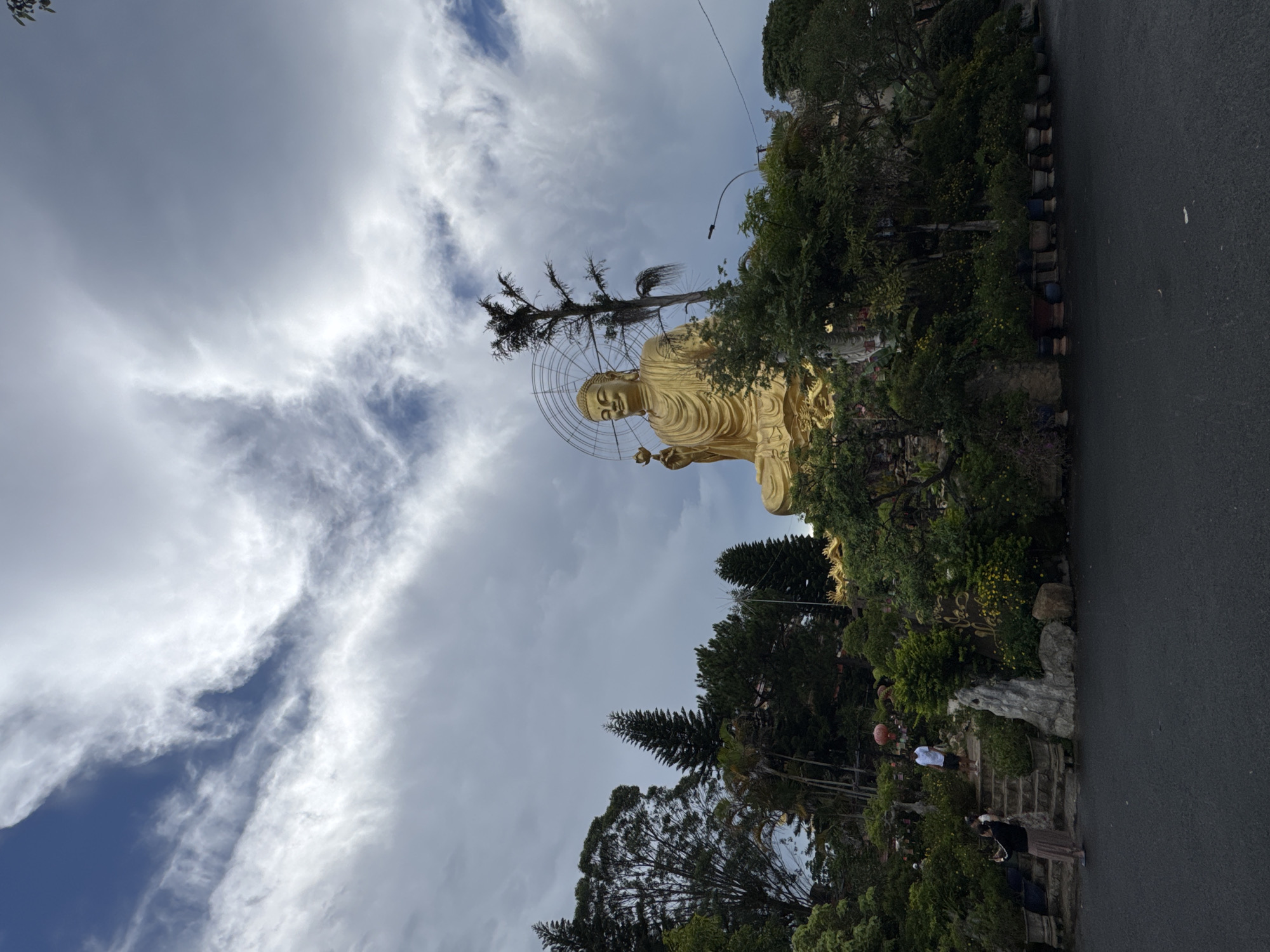 A wide shot captures a colossal golden Buddha statue situated on a hill in Lâm Viên - Đà Lạt, Vietnam. The statue, positioned centrally and dominating the upper half of the frame, is adorned with a halo-like structure and holds a flower in its right hand. The Buddha is surrounded by lush greenery, including numerous trees and potted plants, that cascade down the hillside.
In the foreground, a paved road leads towards the hillside. To the left of the road, a woman in a skirt and top is taking a photograph with her phone, facing the greenery. Further up the steps, a man wearing a white t-shirt and shorts stands looking towards the statue. The sky above is a dramatic mix of blue sky and white, wispy clouds, creating a dynamic atmosphere. The overall impression is one of a serene and majestic religious site set within a natural landscape.