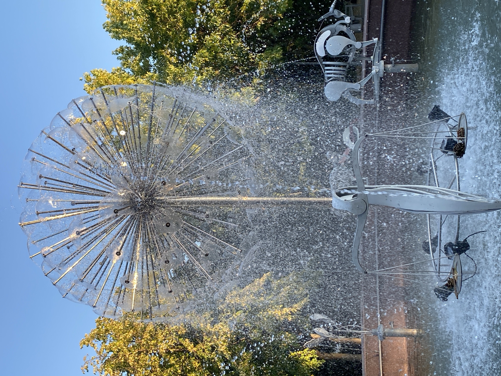 This is a photograph of a fountain in Chișinău, Moldova, specifically located in the Alley of Classics in the Stefan cel Mare Central Park. The fountain is a large, spherical structure made of metal rods, resembling a dandelion or a globe, from which water sprays in a cascading manner. In the foreground, a metal sculpture of a scale, possibly representing justice, is visible, with two pinecones placed on one of its pans. To the right of the scale, a stylized metal sculpture of a bull is mounted. Behind the fountain, lush green trees with yellowing leaves frame the scene, illuminated by the warm light of a clear, blue sky. The overall impression is of a tranquil, artistic public space on a sunny day.